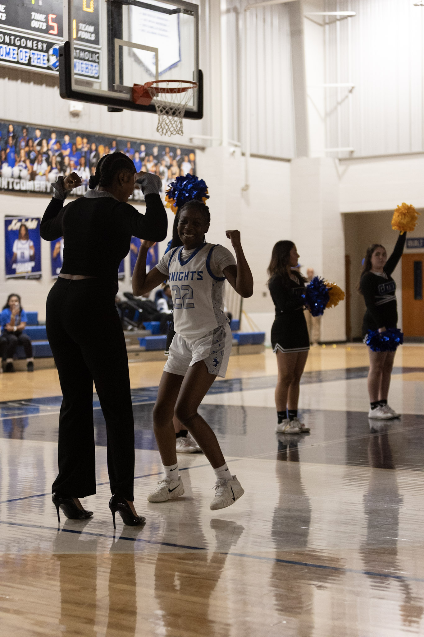 Person in black clothes high-fives girl in basketball uniform during game, cheerleaders with pom-poms in background, gymnasium setting