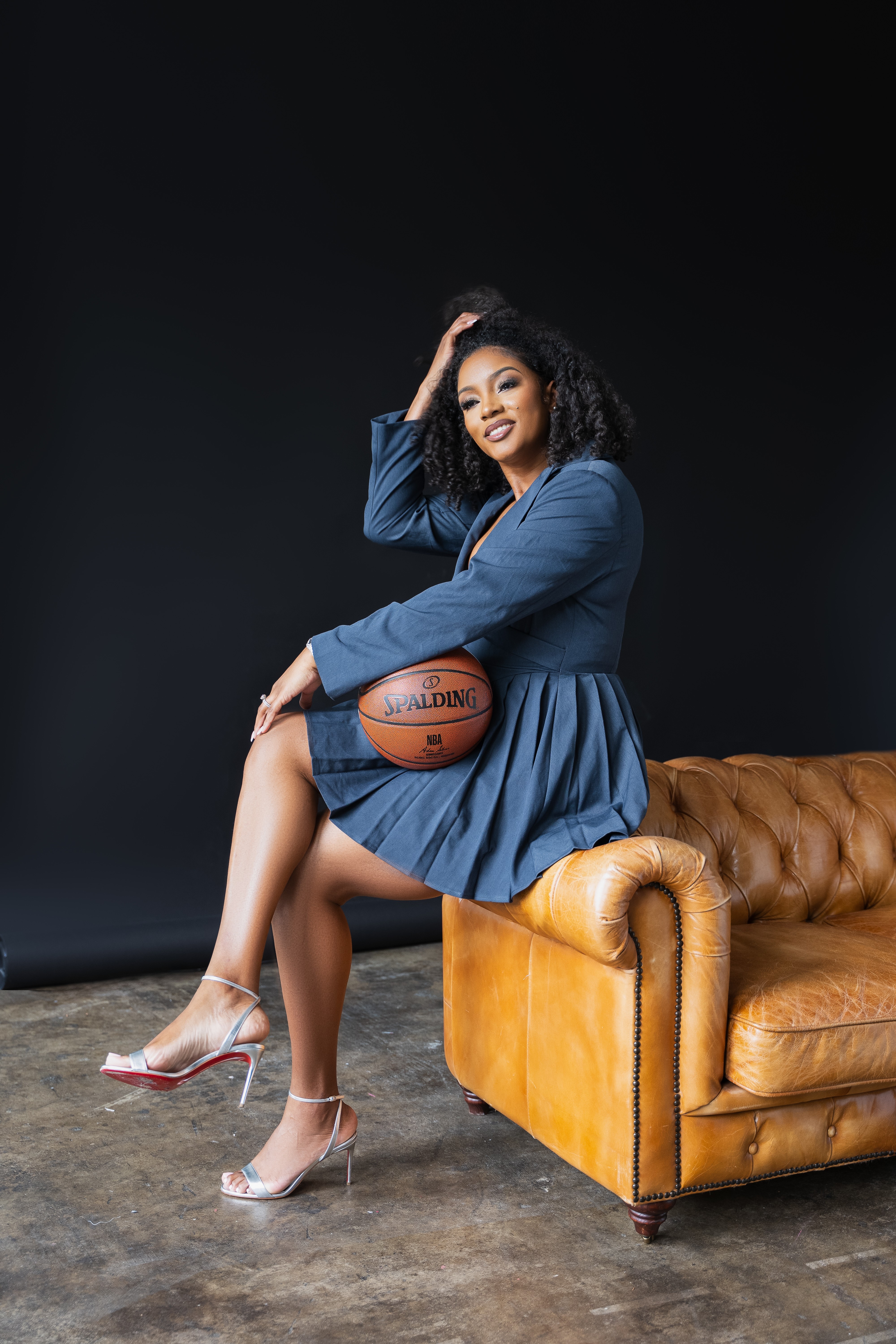 Woman in blue dress sitting on a brown leather sofa holding a basketball, with a dark background.
