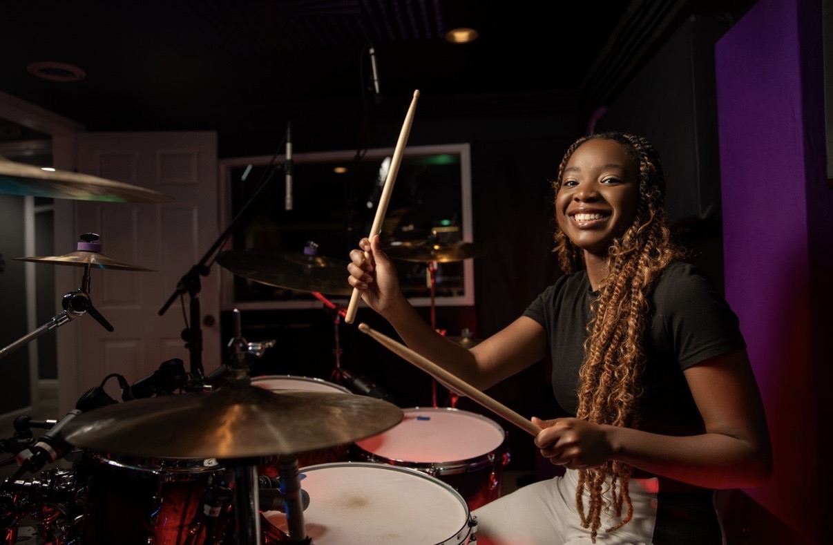 Young woman smiling while playing drums in a music studio, holding drumsticks, with a purple wall behind her.