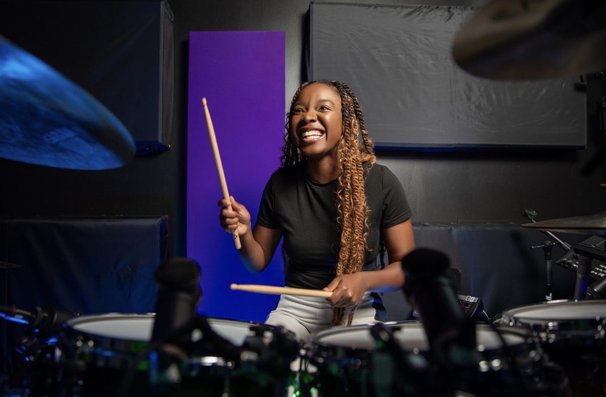 Woman smiling while playing drums, holding drumsticks, in a music studio with dark background and purple panel.