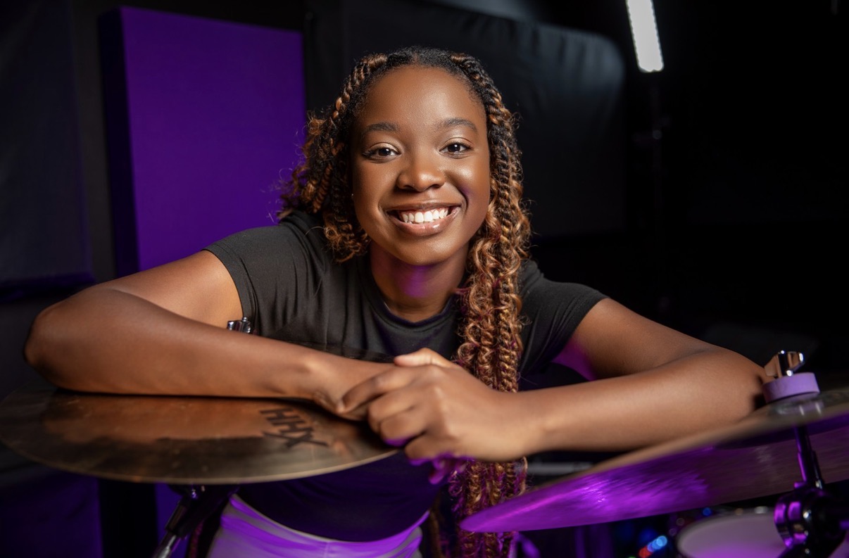 Smiling girl with curly hair resting her arms on a drum set in a dark studio with purple lighting.