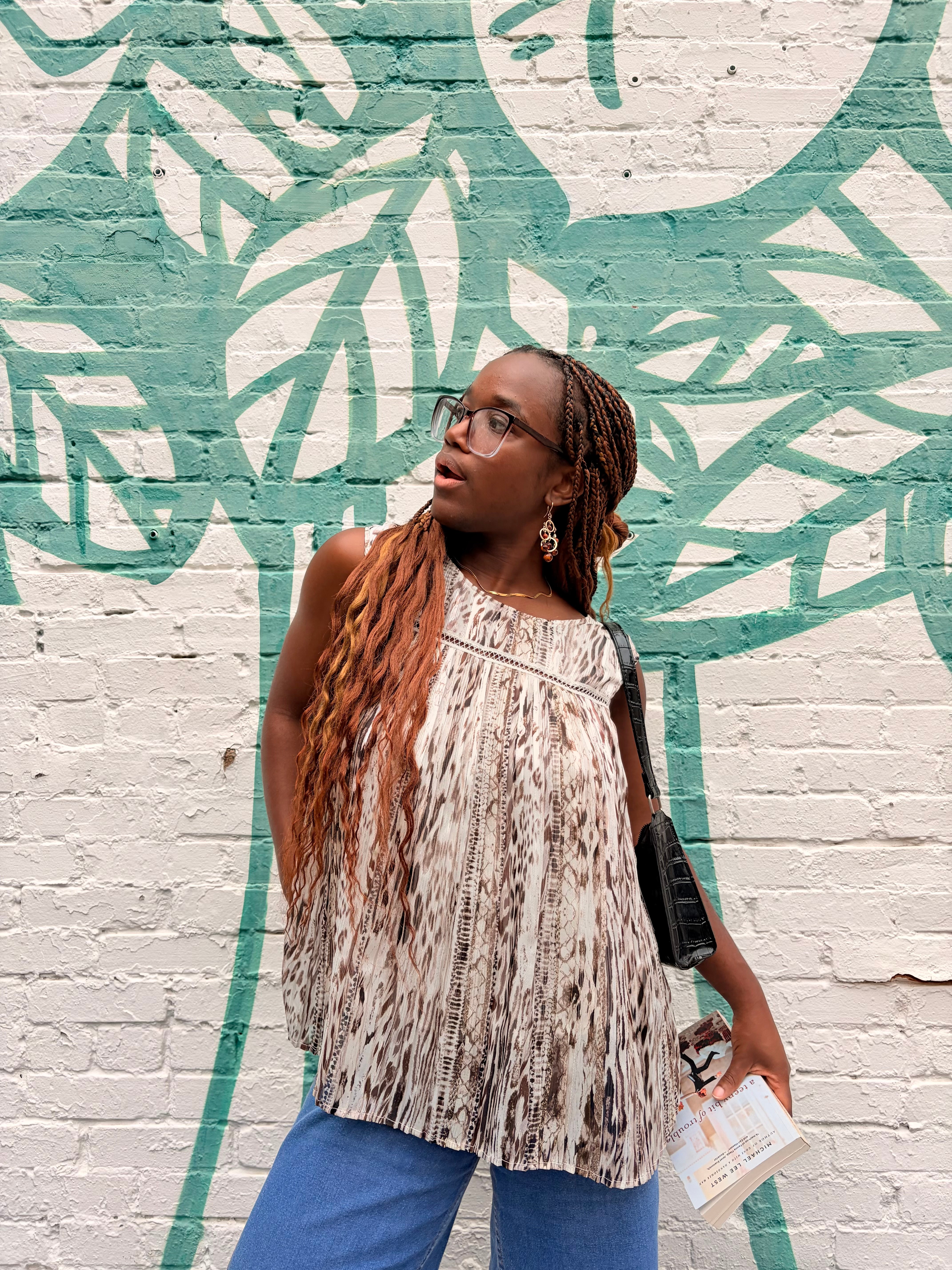Woman with glasses and long braided hair stands in front of a mural with leaf patterns on a brick wall.