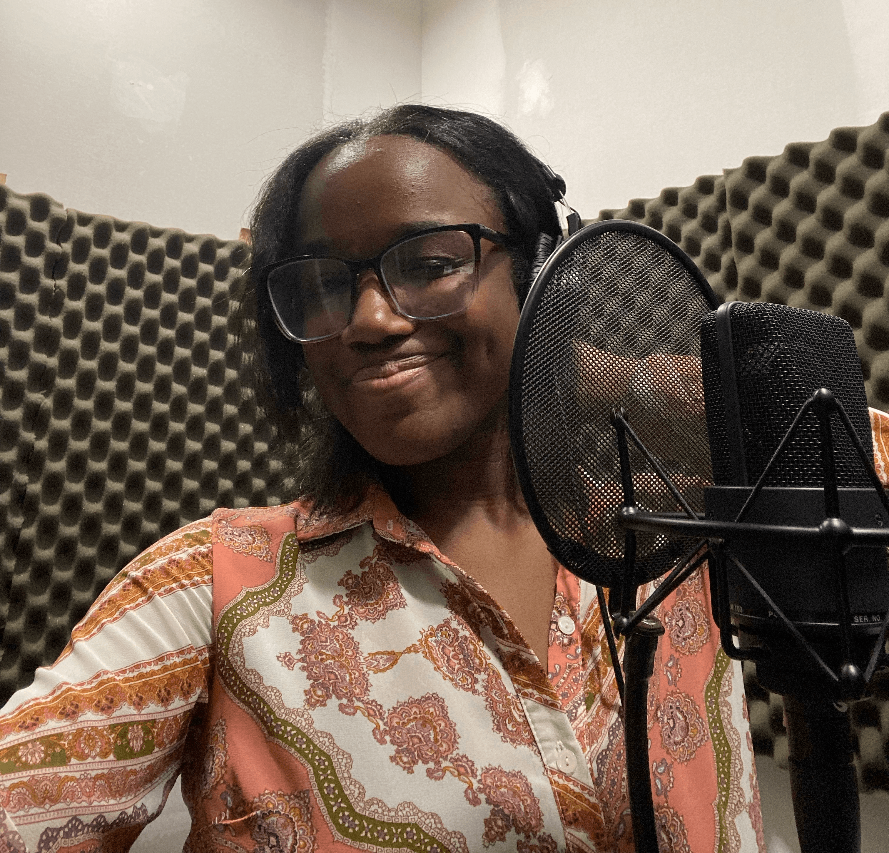 Woman with glasses smiling in a recording studio with soundproof foam panels and a microphone.