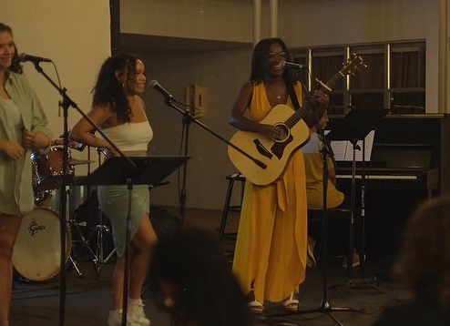 Group of women performing music on stage with instruments, including guitar and drums, in an indoor setting.