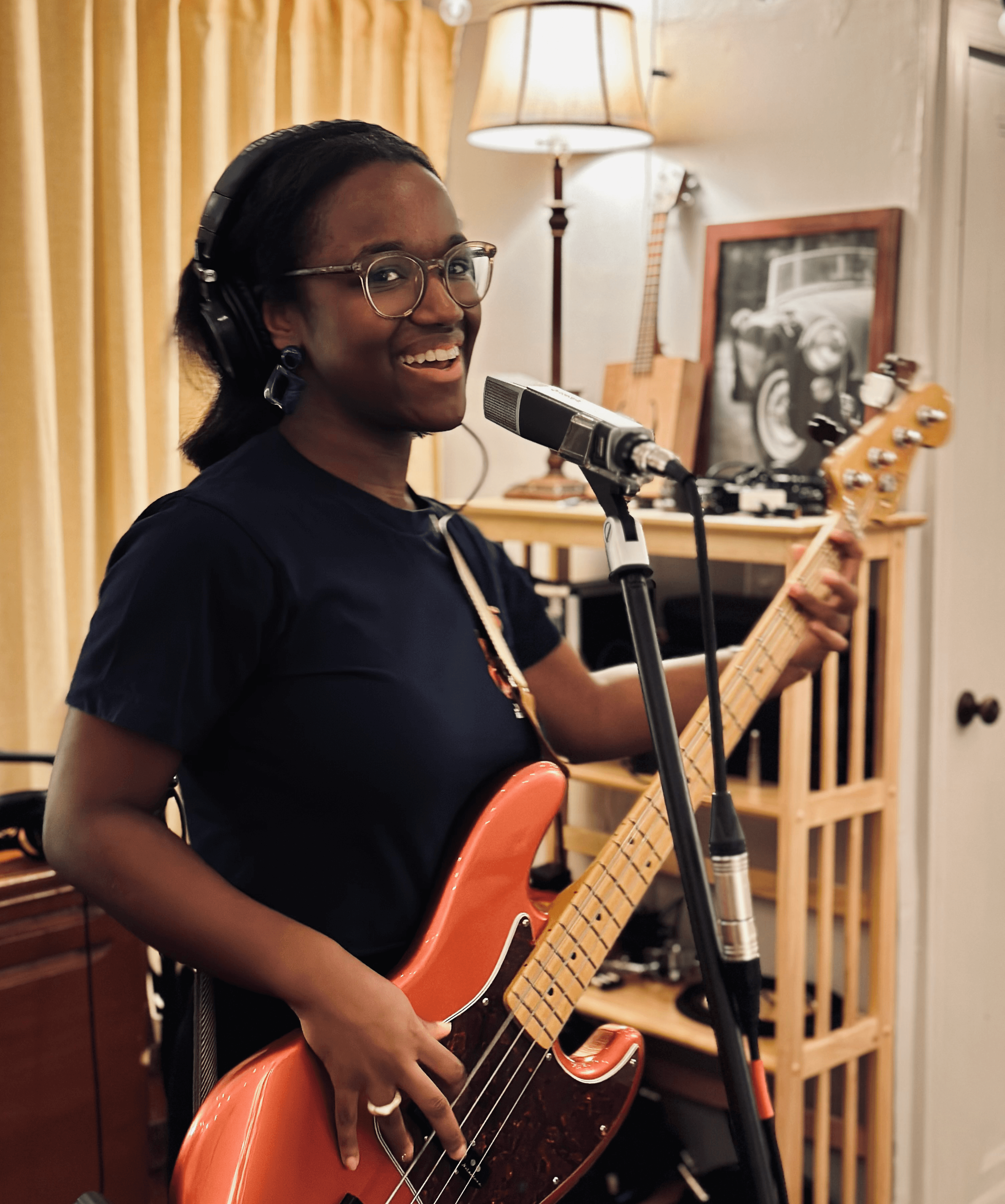Woman with glasses and headphones playing an electric guitar in a room with a lamp and framed picture.