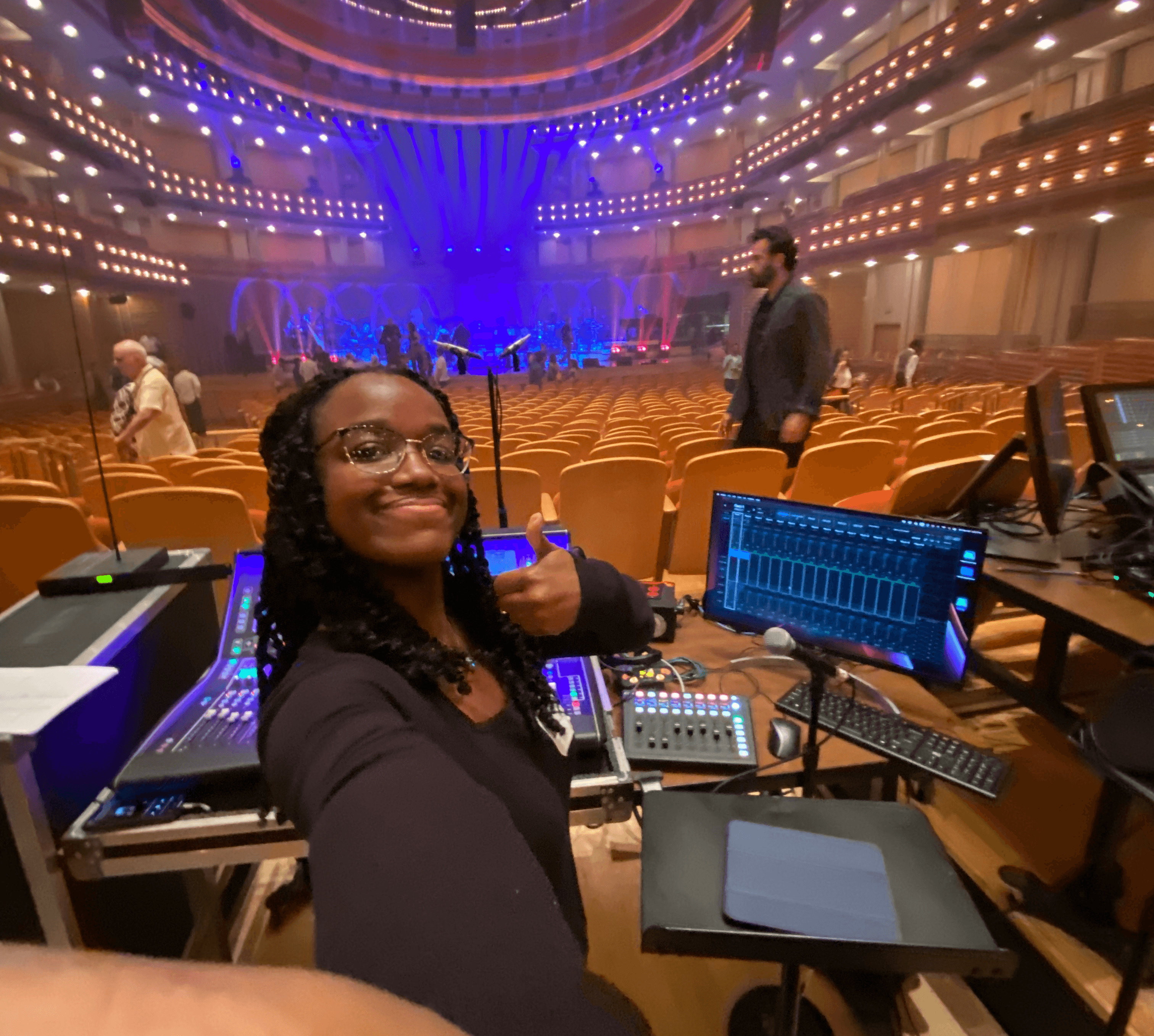 Woman smiling and giving a thumbs-up in a large auditorium with stage and seating, surrounded by audio equipment and monitors.