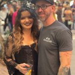 A smiling woman with long wavy hair and dark lipstick holds a drink, standing next to a man with glasses and a cap.