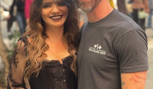 A smiling woman with long wavy hair and dark lipstick holds a drink, standing next to a man with glasses and a cap.