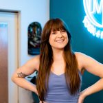 Young woman with long brown hair smiling, standing indoors near a neon sign, hands on hips, wearing a sleeveless top.