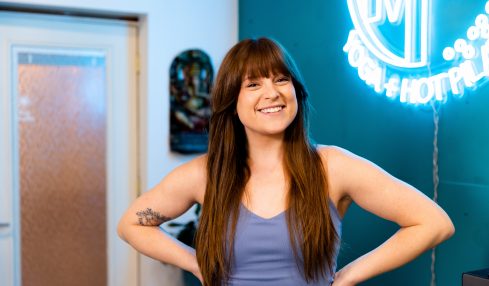 Young woman with long brown hair smiling, standing indoors near a neon sign, hands on hips, wearing a sleeveless top.