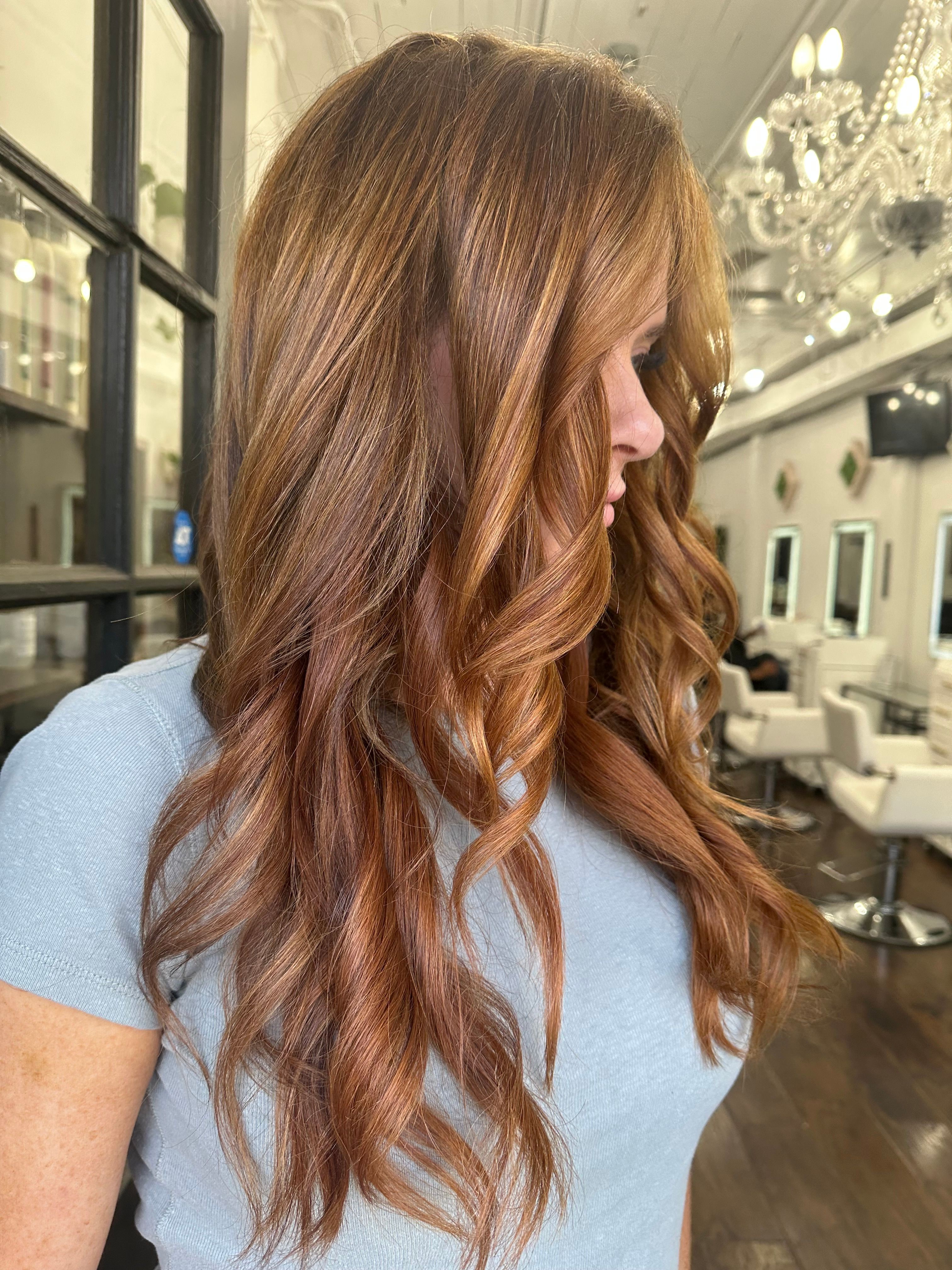Side view of a woman with long, wavy, reddish-brown hair in a well-lit room with elegant decor.