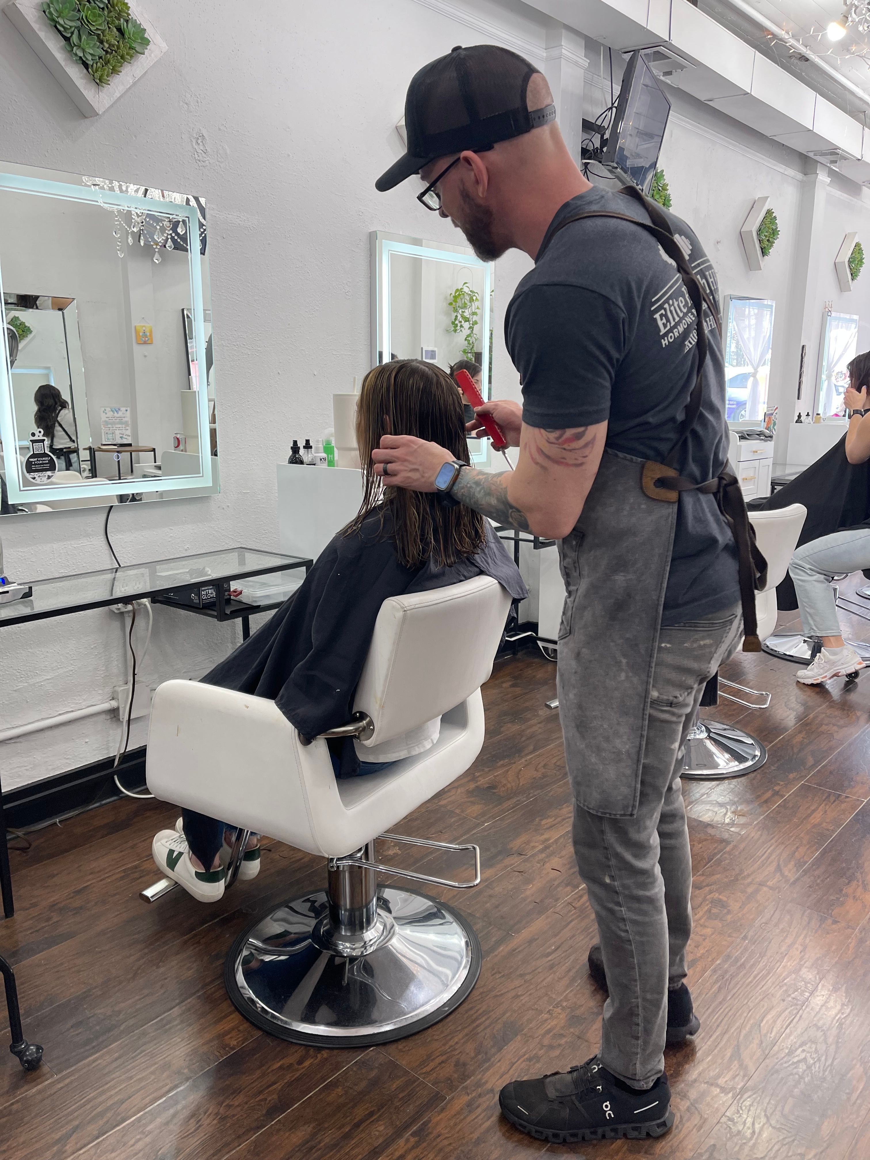 Person in a salon chair with a cape, receiving a haircut, in a modern salon with large windows and wooden floor.