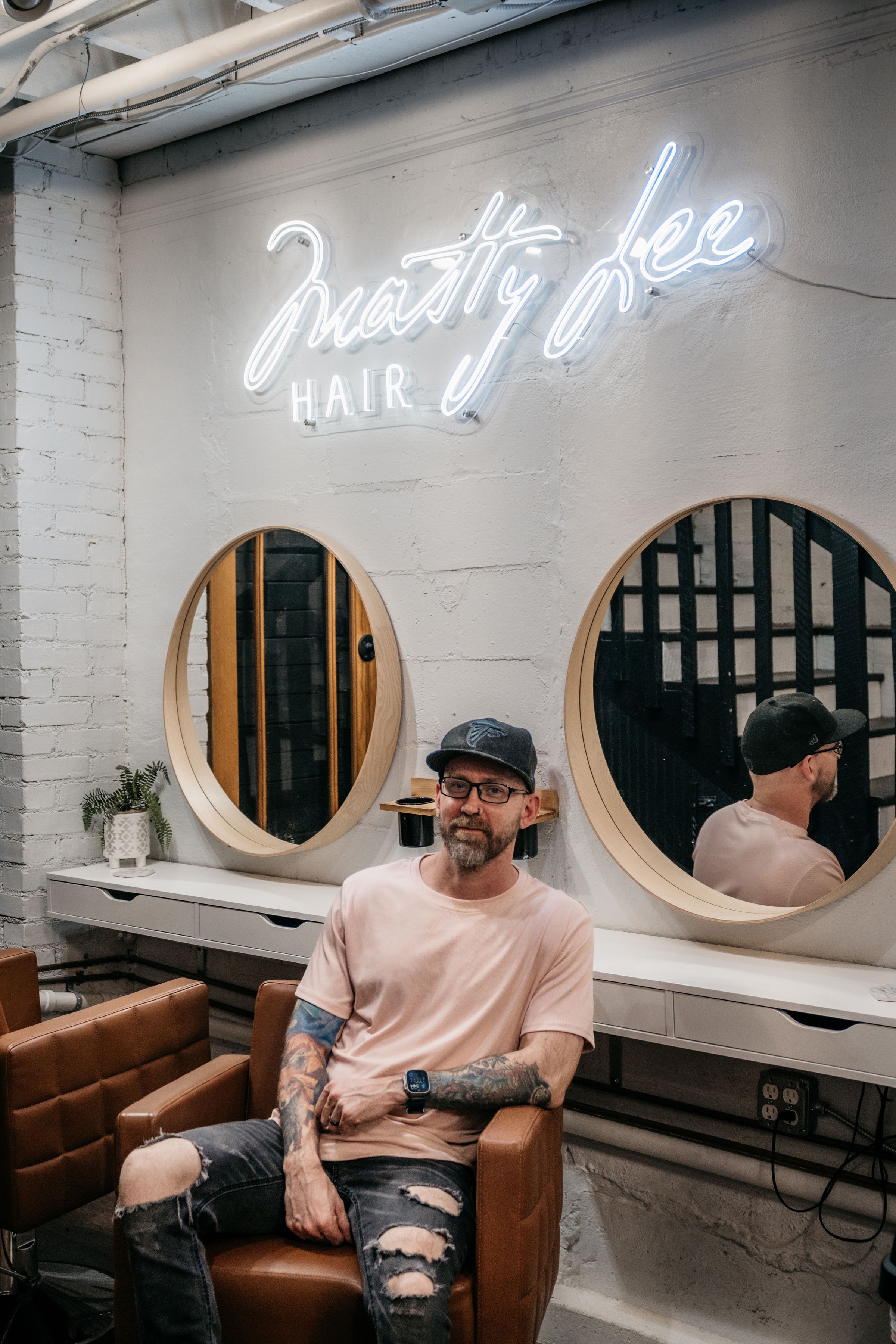 Man with tattoos and glasses sits in a hair salon, mirrors and neon sign in background.