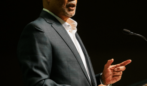 Man in suit speaking at a podium with a microphone, gesturing with his right hand, dark background