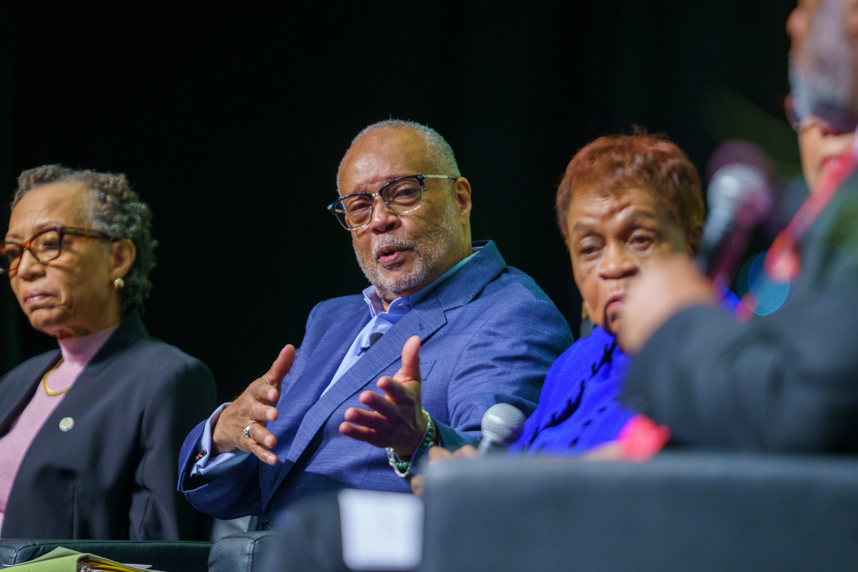 Four people seated at a panel discussion, with a dark background, one speaking and gesturing with hands.