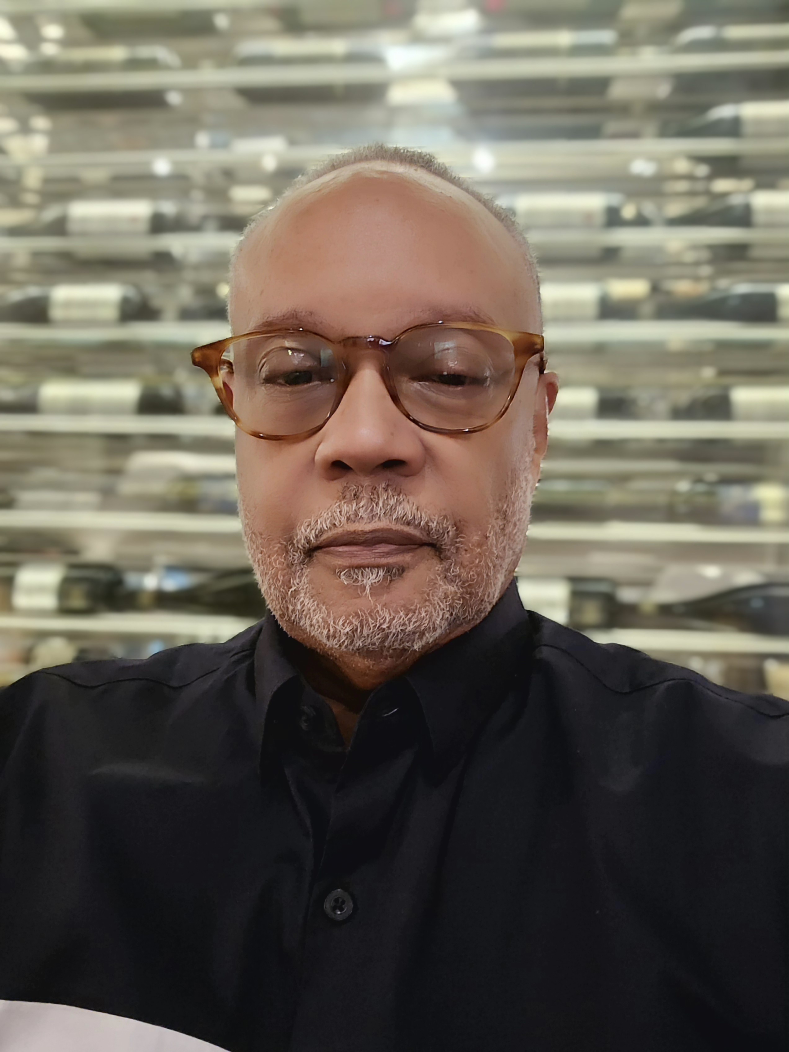 A man with glasses and a beard wearing a black shirt, standing in front of a blurred background.