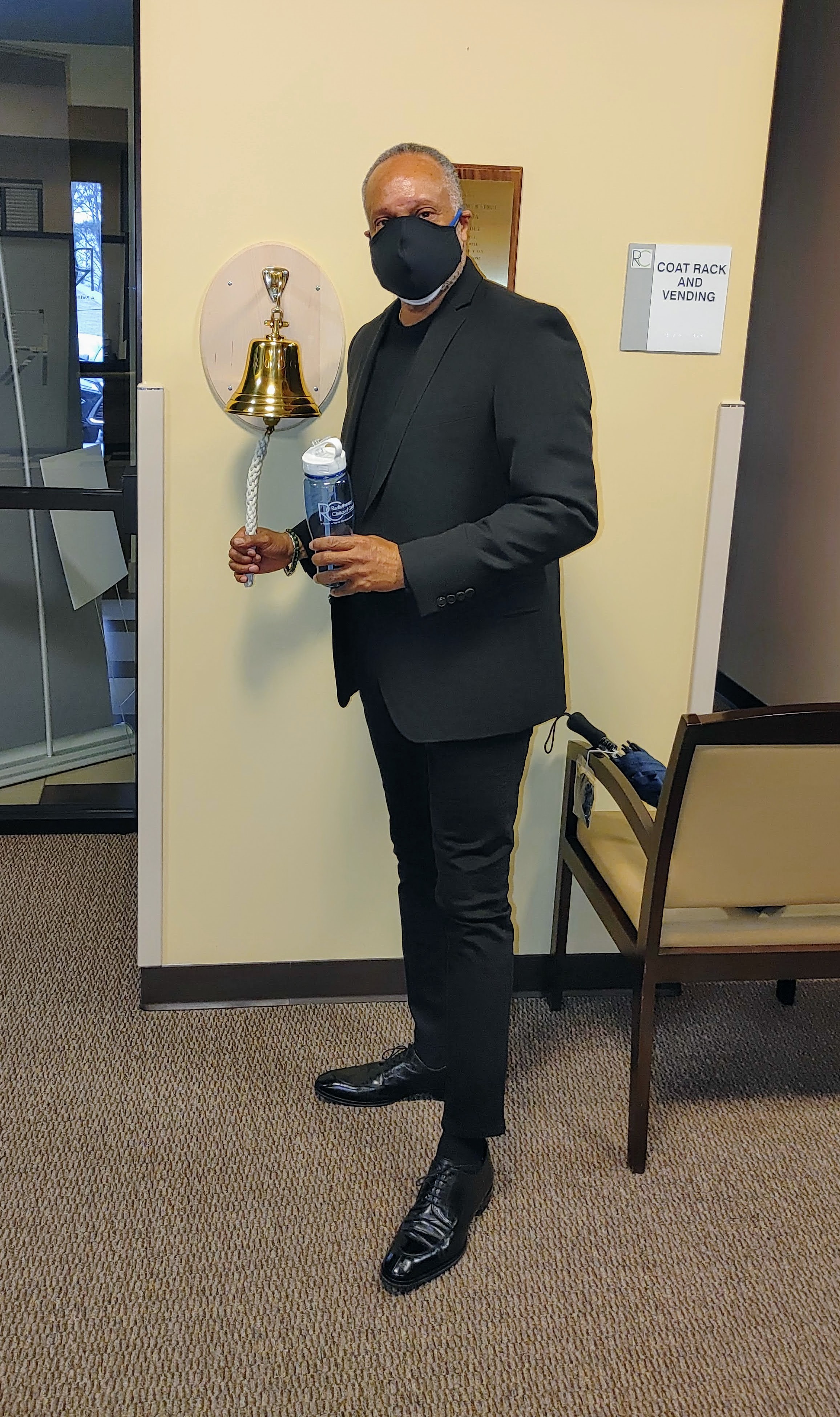 Man in black suit and mask holding water bottle and glass, standing indoors near a wall and a chair.