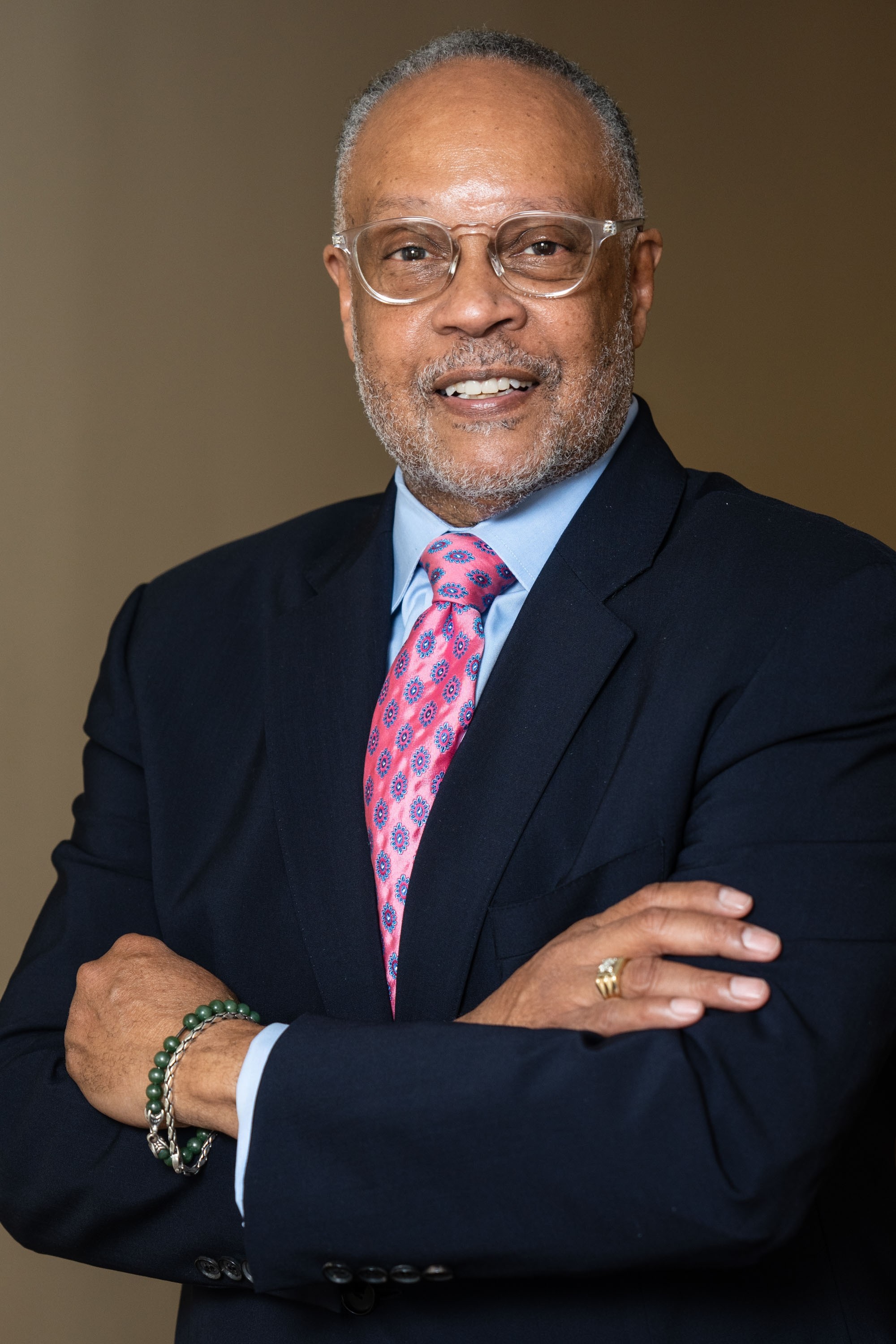 Portrait of an older man with glasses, gray beard, wearing a dark suit, pink patterned tie, and a bracelet, smiling with arms crossed.