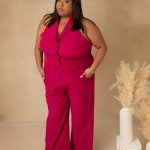 Woman in pink sleeveless jumpsuit with a headband, standing with hands in pockets, neutral background, decorative pampas grass, and vase.
