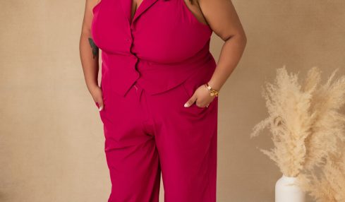 Woman in pink sleeveless jumpsuit with a headband, standing with hands in pockets, neutral background, decorative pampas grass, and vase.