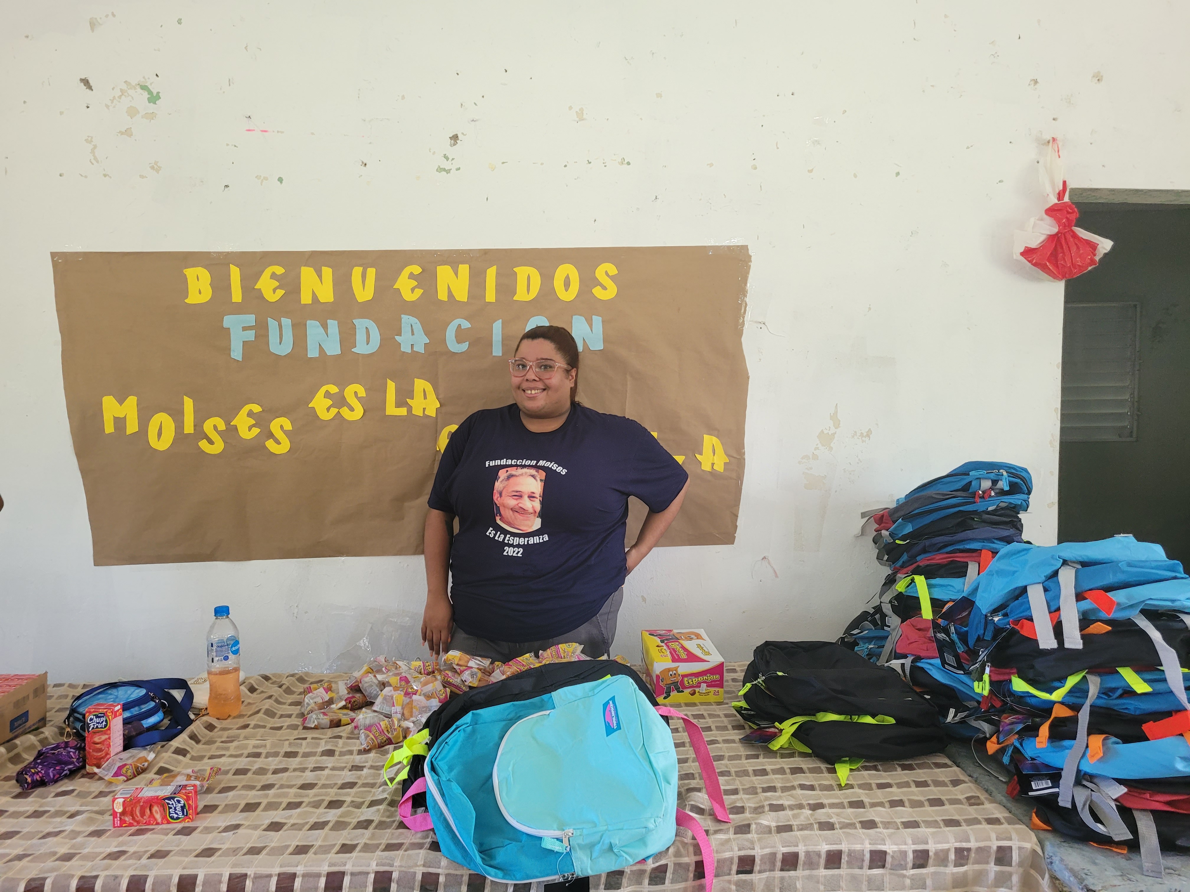 Person standing in front of a welcome sign, table with backpacks and supplies, wall with hanging decoration, indoor setting.