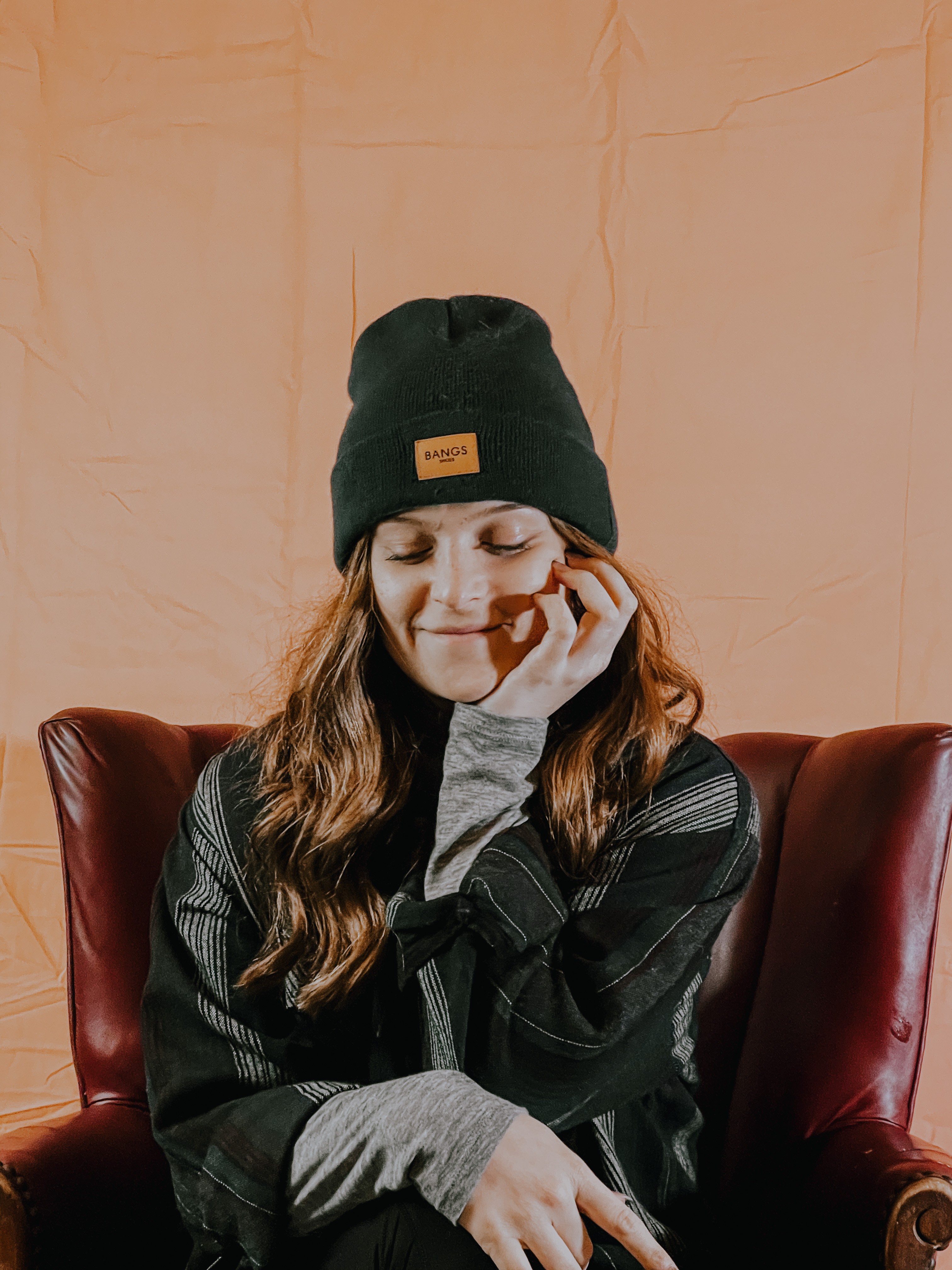 Young woman with long hair wearing a black beanie, sitting on a red chair, smiling with eyes closed, hand near face.