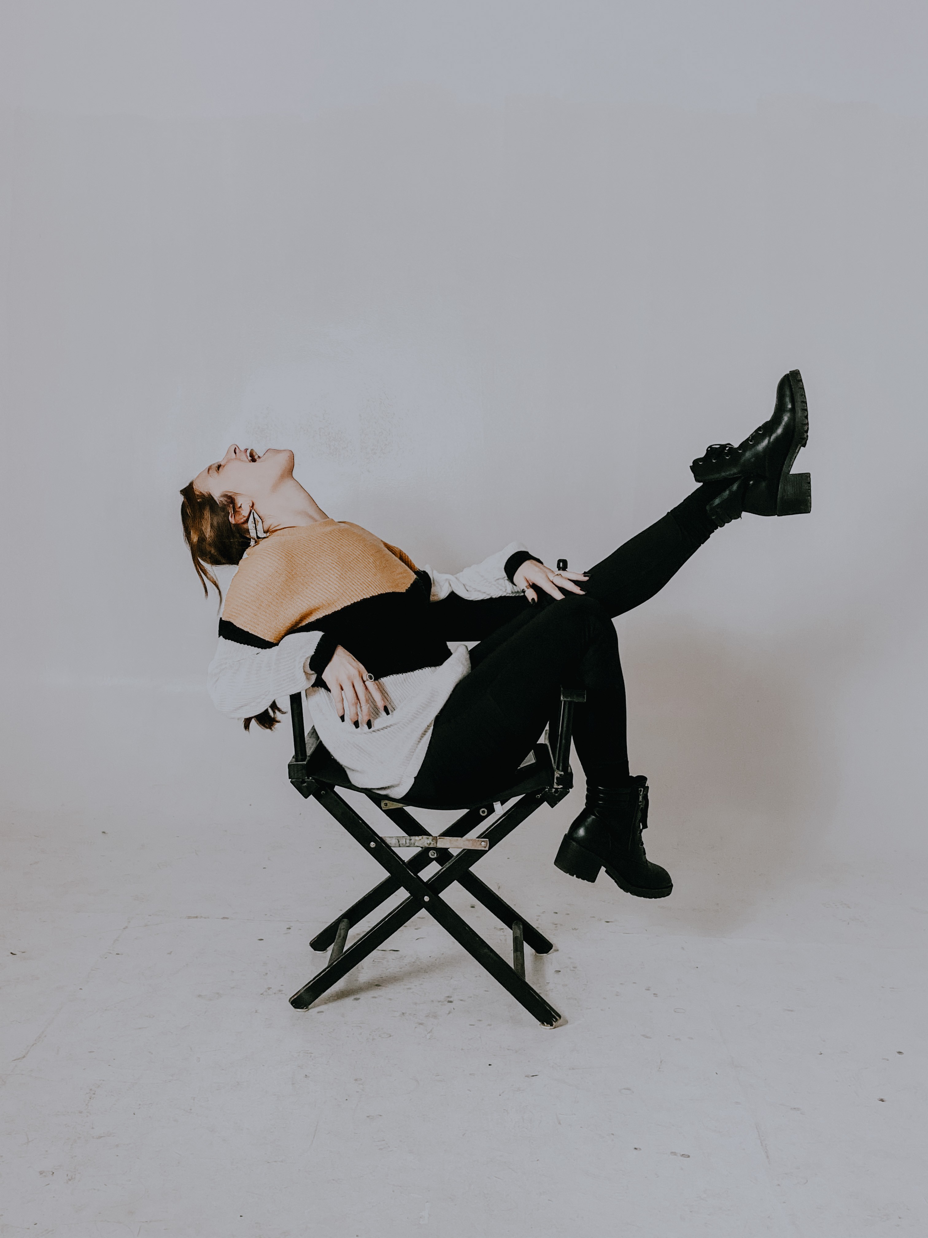 Person sitting on a folding chair with legs extended, head tilted back, in a minimal studio setting.