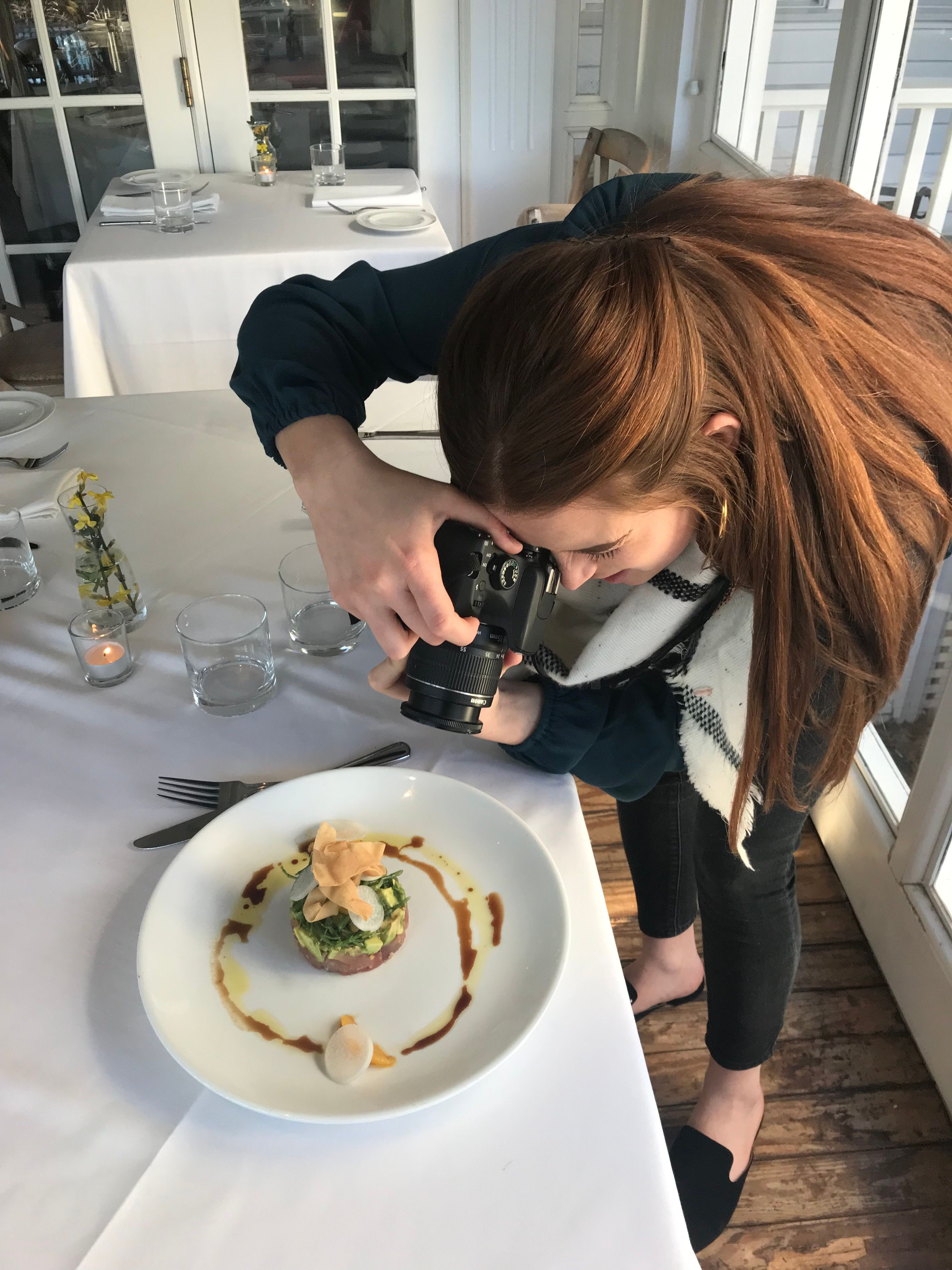 Person with long hair taking a photo of a plated dessert at a table in a bright room.
