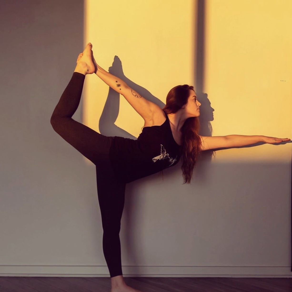 Person performing a yoga pose against a wall, with shadow cast on the wall, in a room with warm lighting.