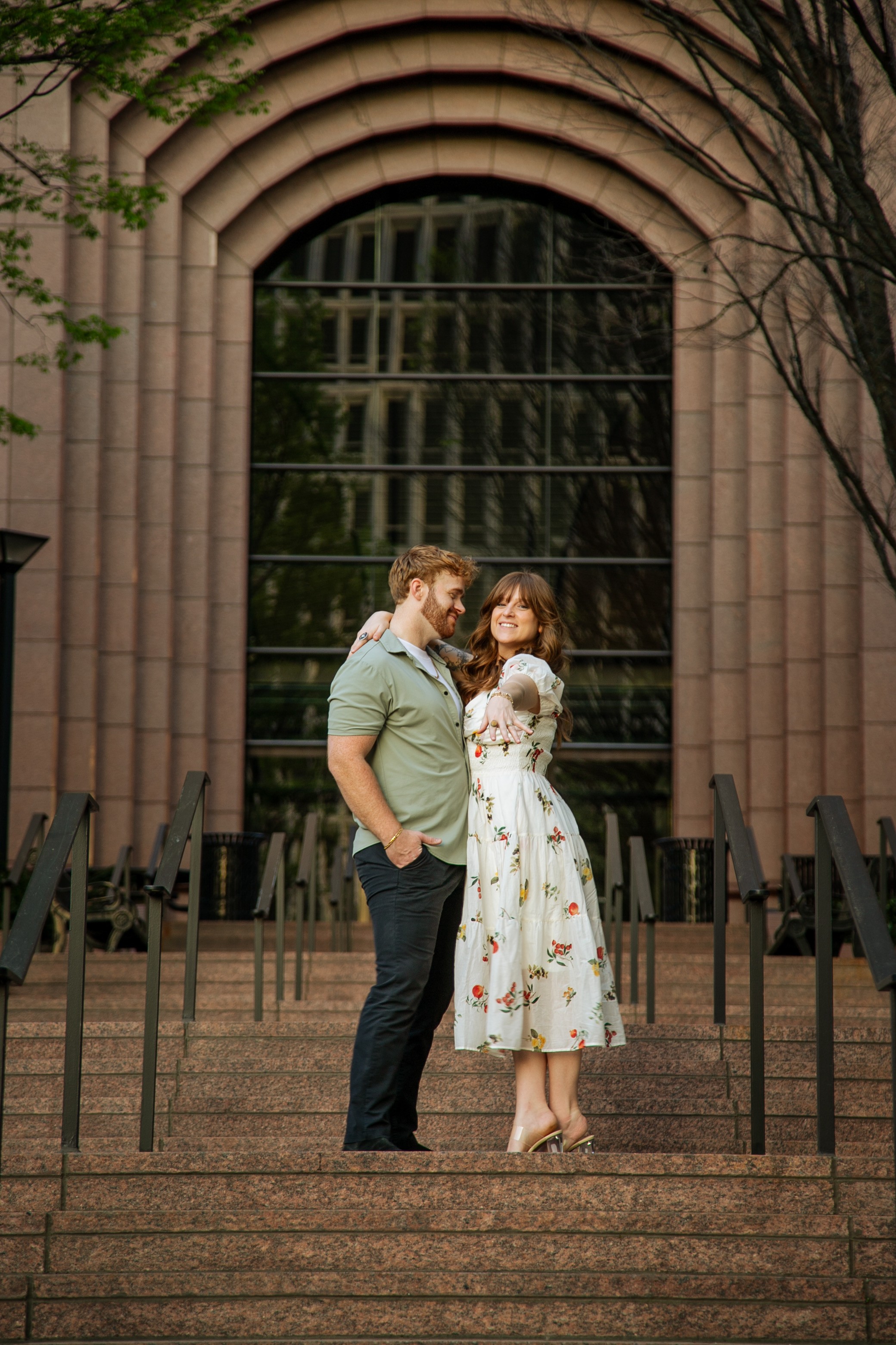 A man and woman stand close together on steps in front of a large arched window, holding hands and looking at each other.