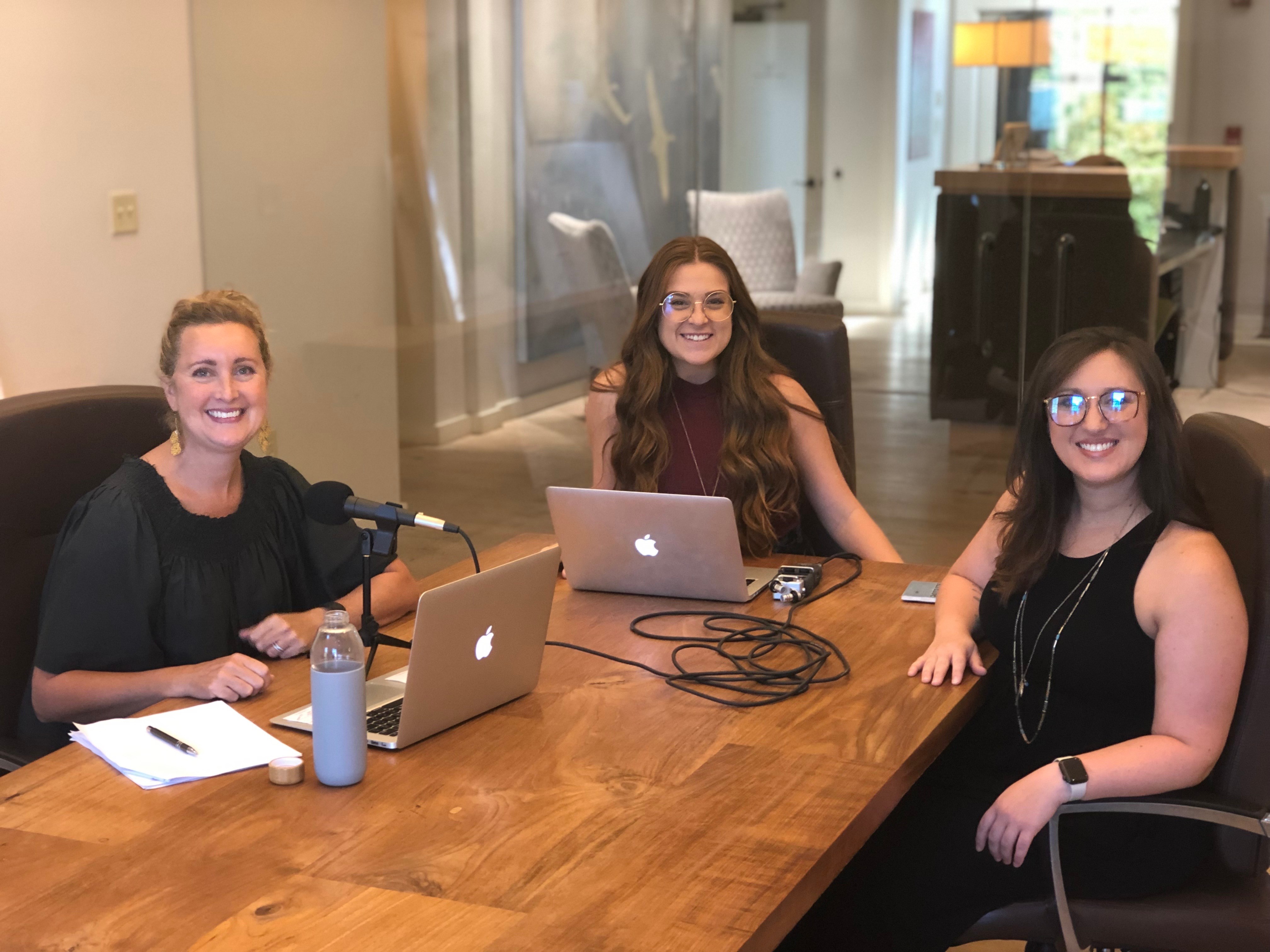 Three women sitting at a wooden table with laptops, smiling in a room with large windows and a piano in the background.
