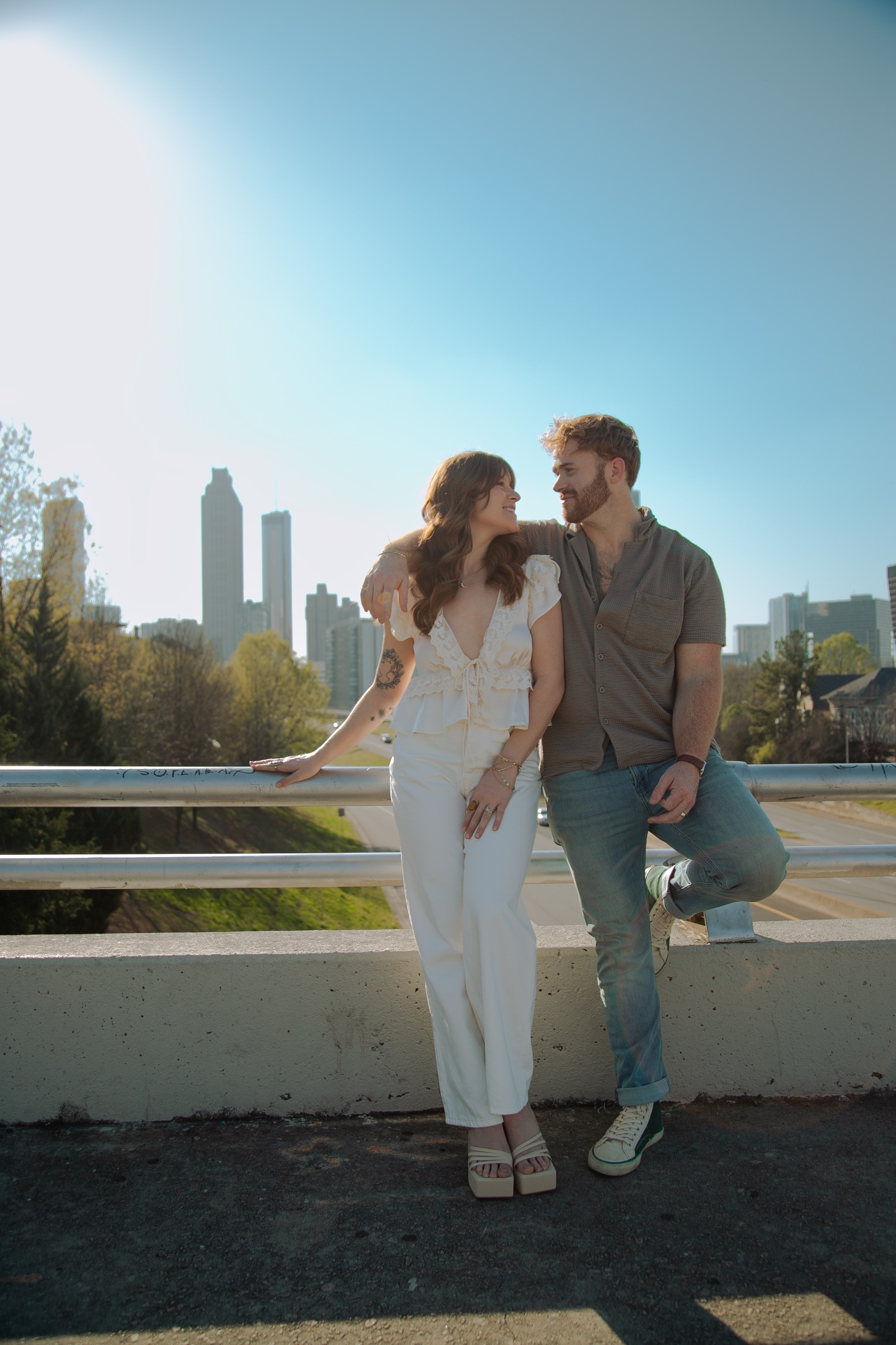 A couple stands on a bridge with a city skyline in the background, holding hands and looking at each other.
