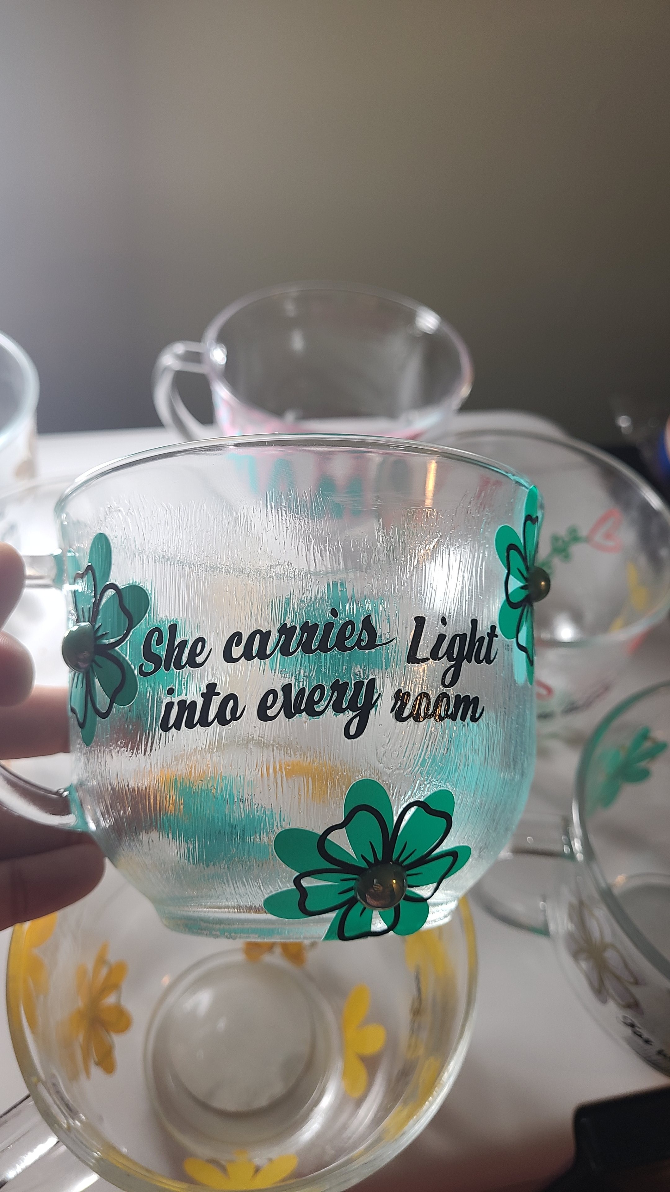 Clear glass cup with teal and yellow floral design and text, placed on a table with other cups in the background.