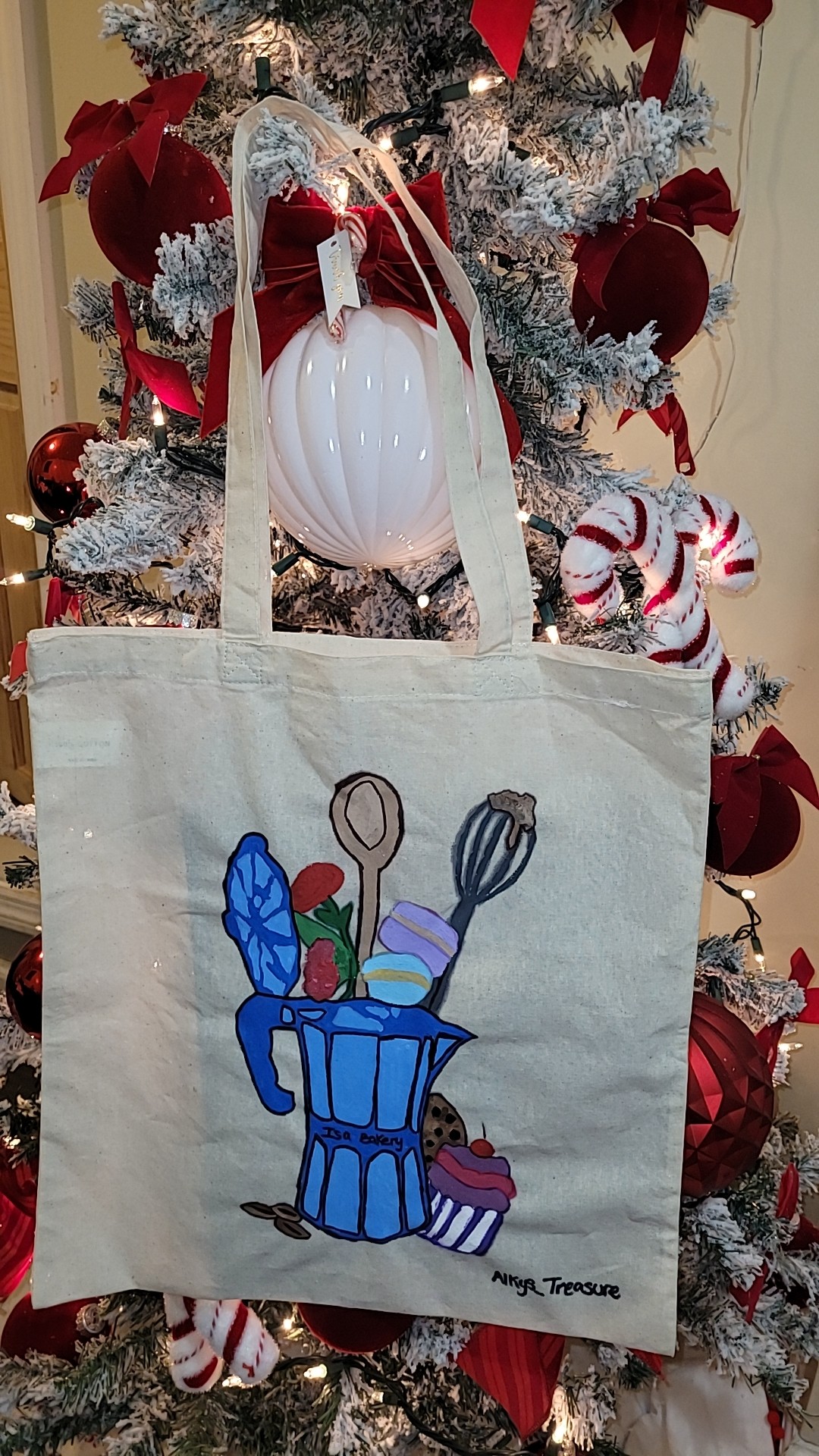 Canvas tote bag with a colorful illustration of kitchen utensils in a blue container, hanging in front of a decorated Christmas tree.