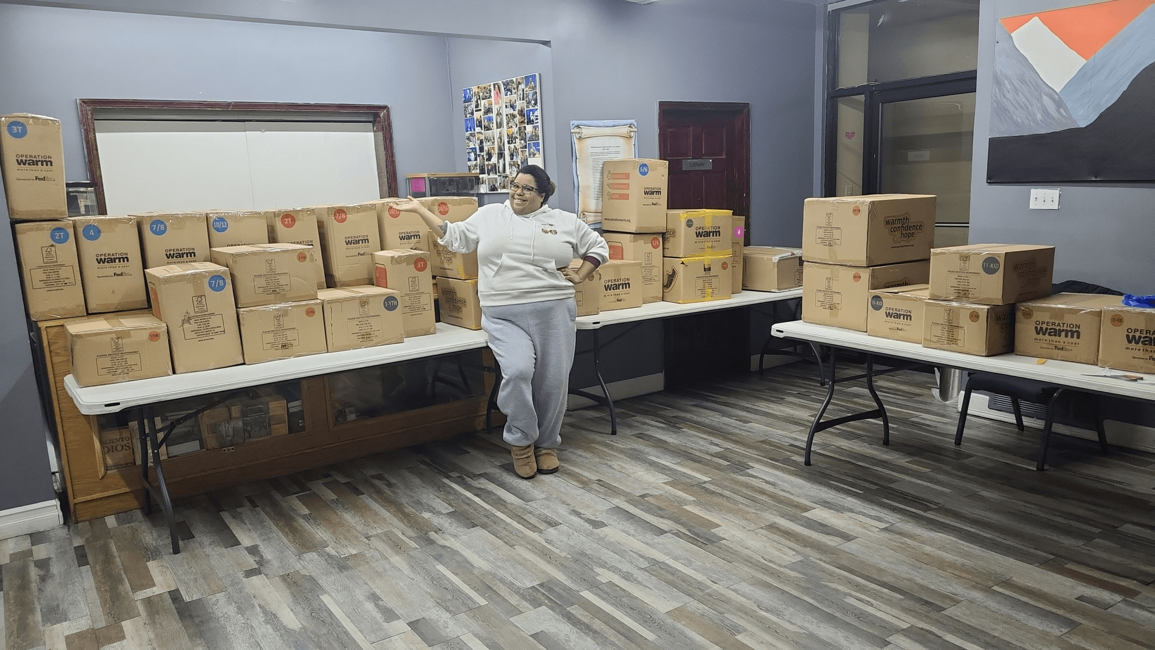 Person standing in a room with stacked boxes on tables and shelves, gray walls, and a window.