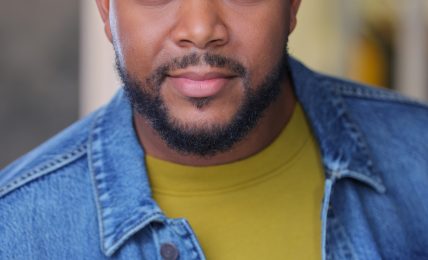 Portrait of a man with a beard and short hair, wearing a denim jacket and yellow shirt, looking at the camera.