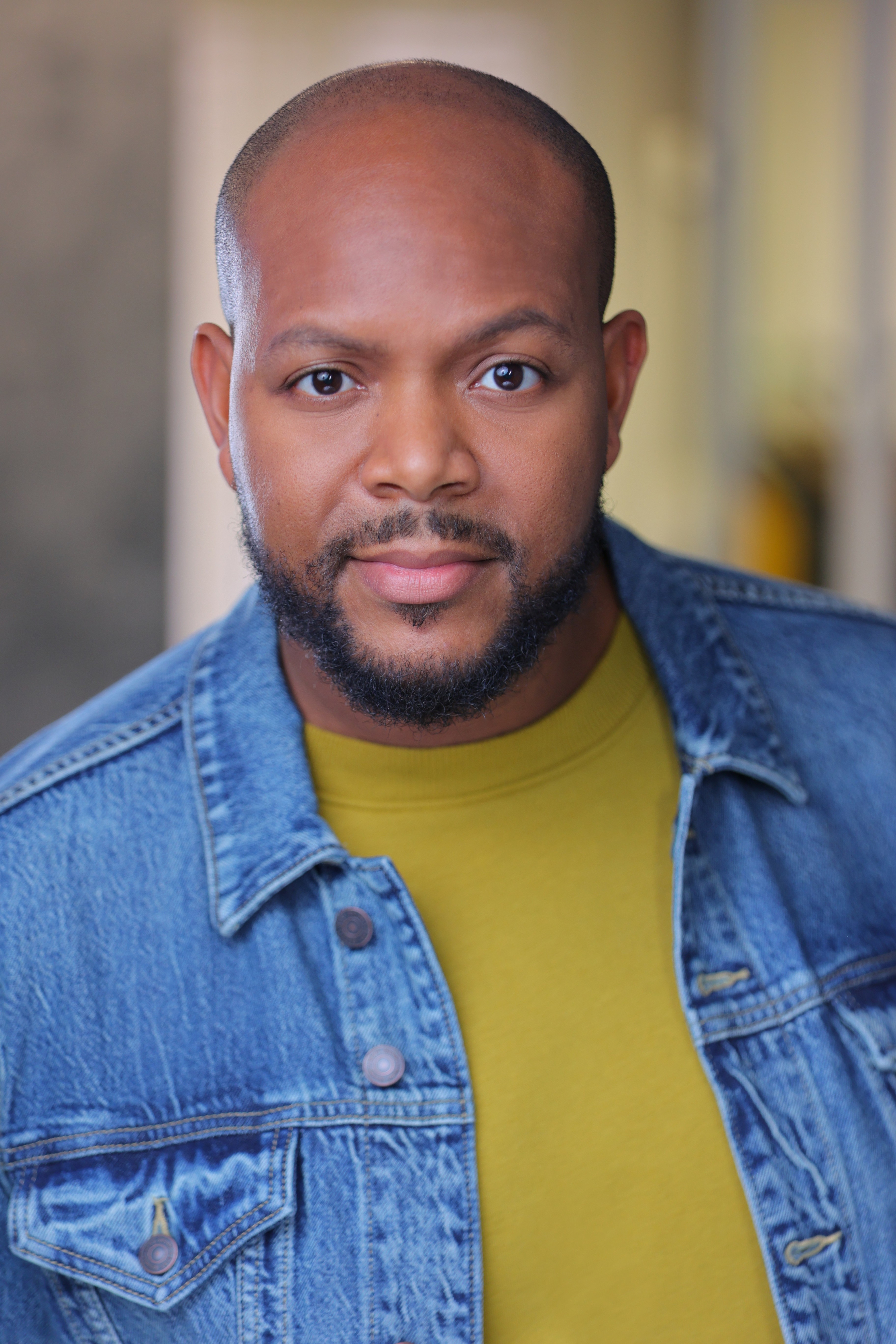 Portrait of a man with a beard and short hair, wearing a denim jacket and yellow shirt, looking at the camera.