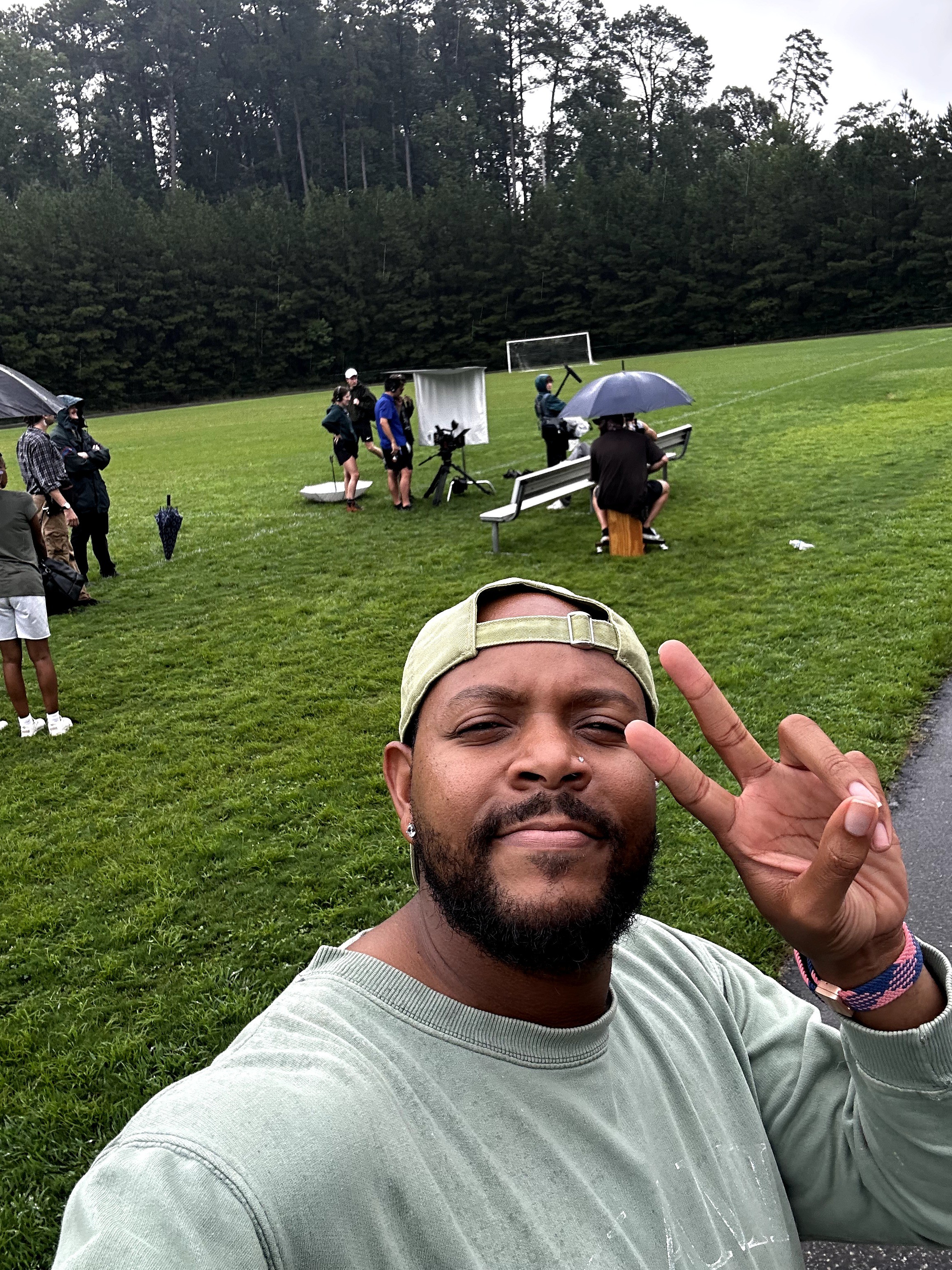 Man taking selfie on a golf course with people and golf equipment in the background, holding up a peace sign.