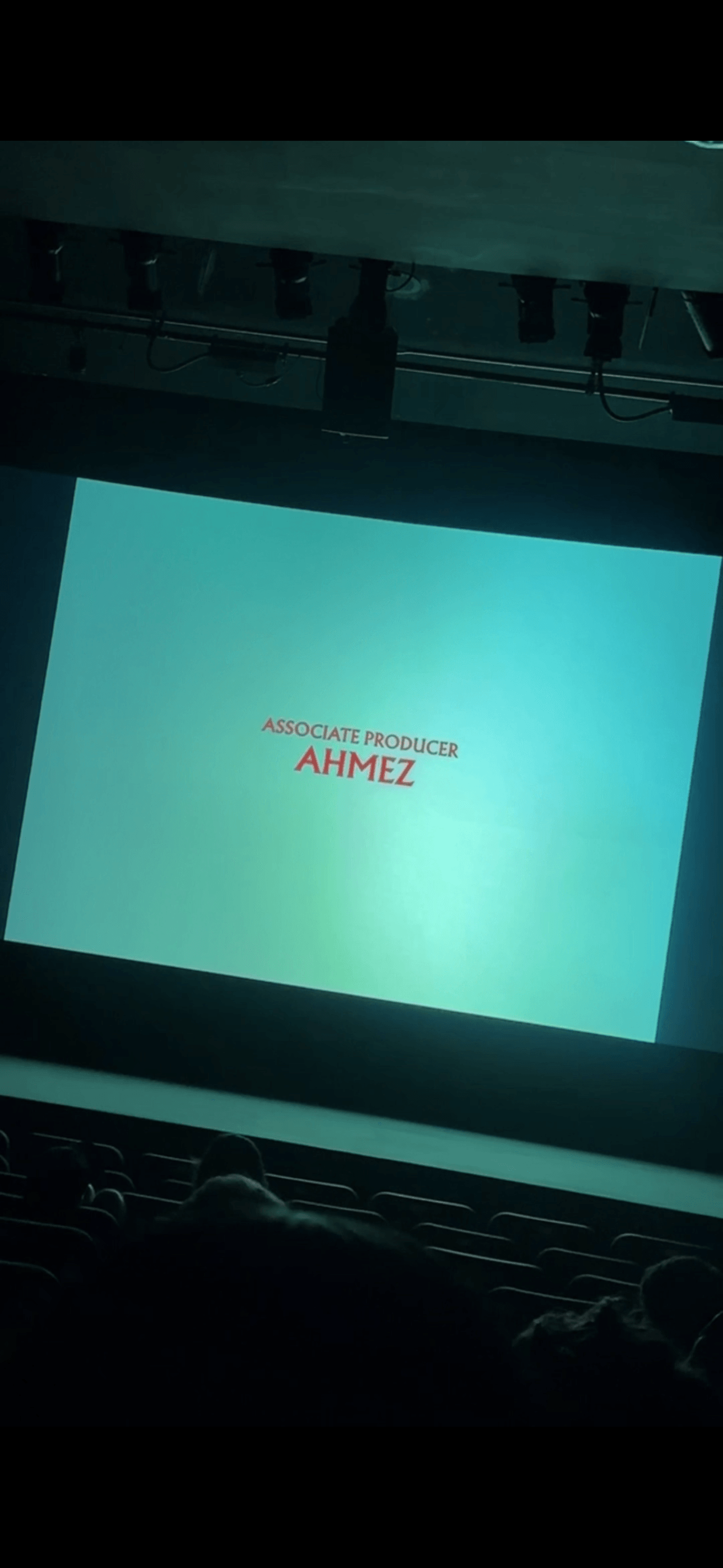 Projection screen displaying text, with a dark theater setting and visible ceiling equipment above.