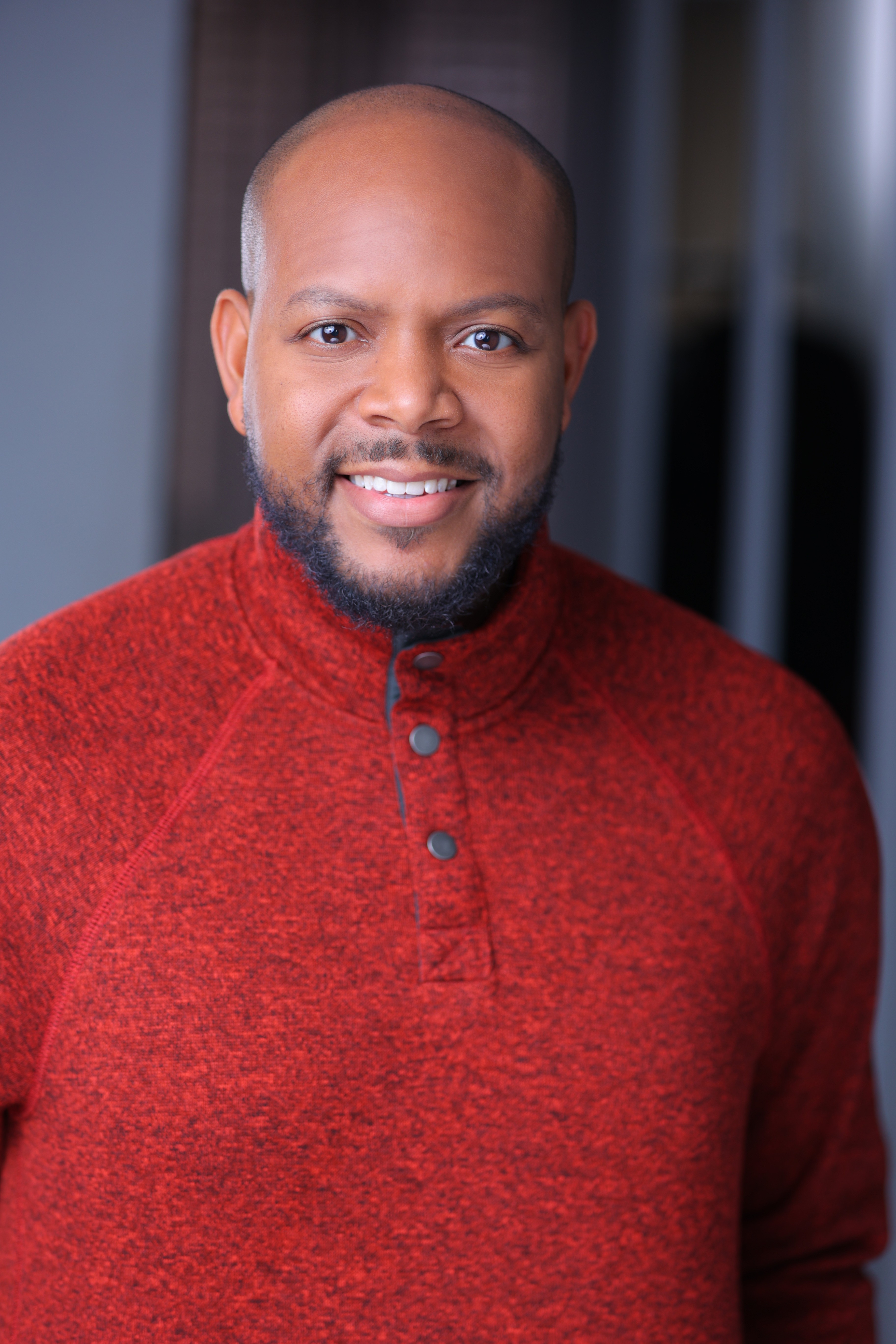 Man with a beard and a shaved head wearing a red sweater, smiling, indoors with blurred background.