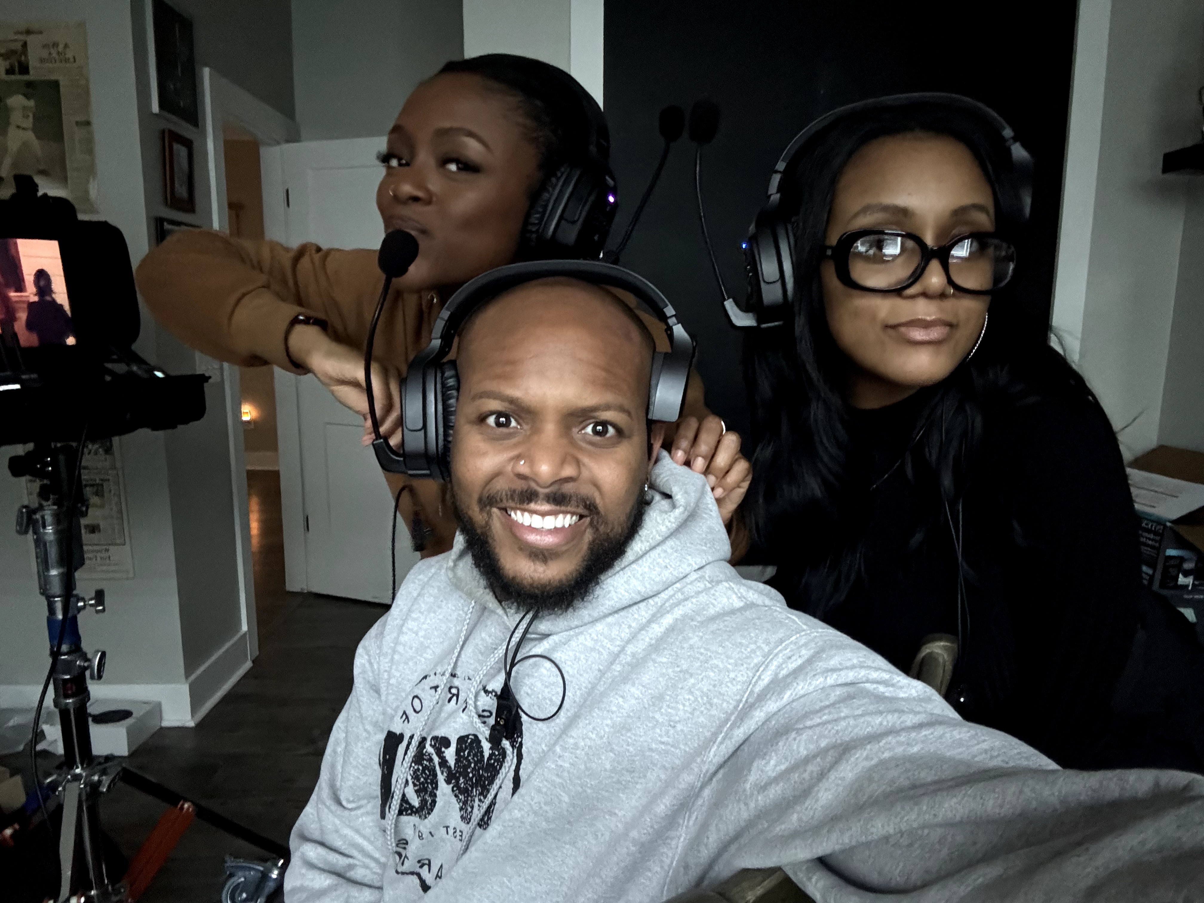 Three people wearing headphones, smiling and posing for a photo indoors, with a camera on a tripod nearby.