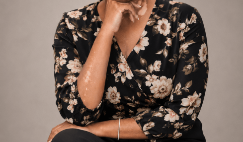 Woman sitting on a stool, wearing a floral blouse and black pants, smiling with her hand near her chin.