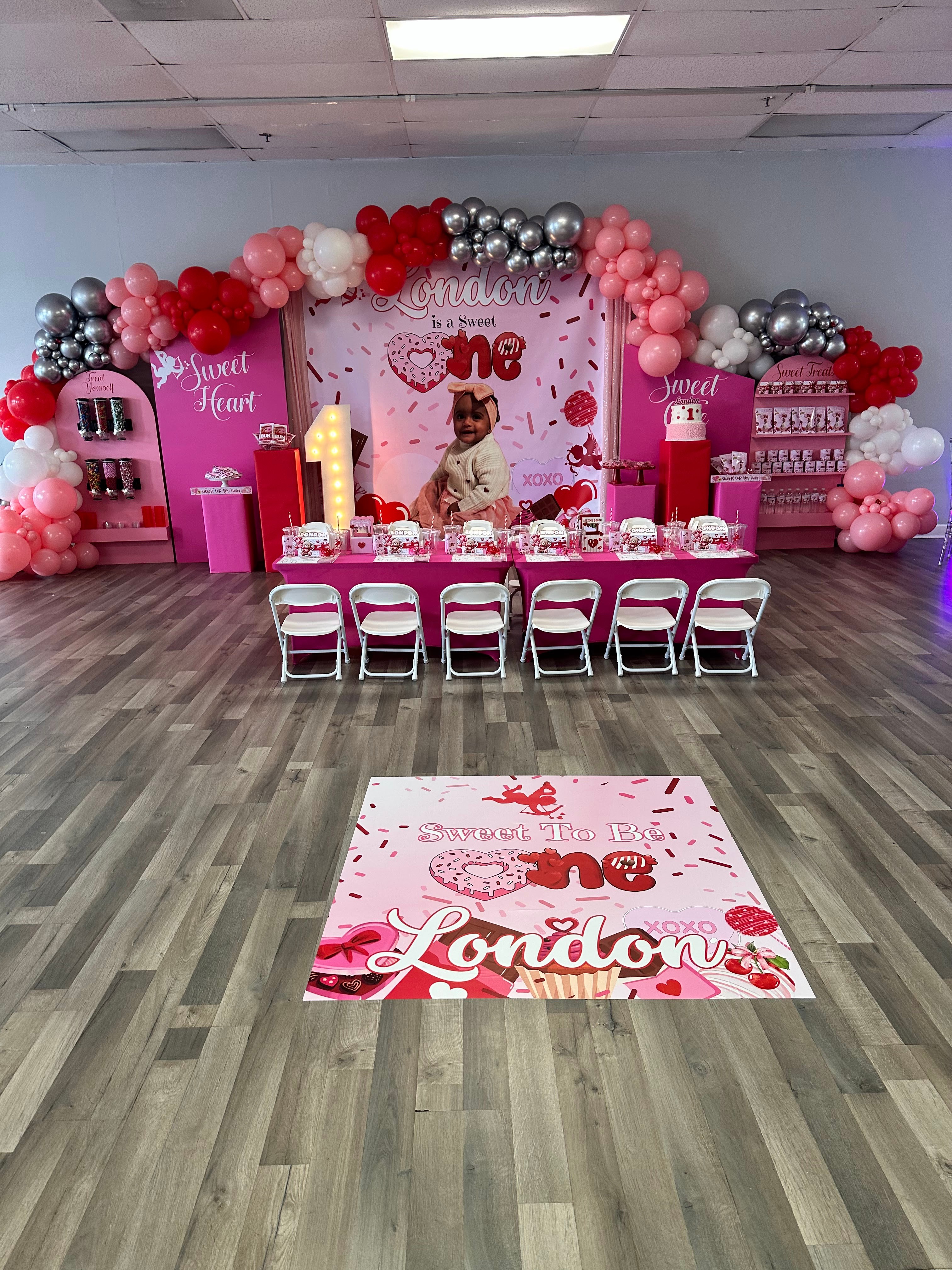 Party setup with pink, red, and silver balloons, a decorated backdrop, and a table with chairs for a child's celebration.