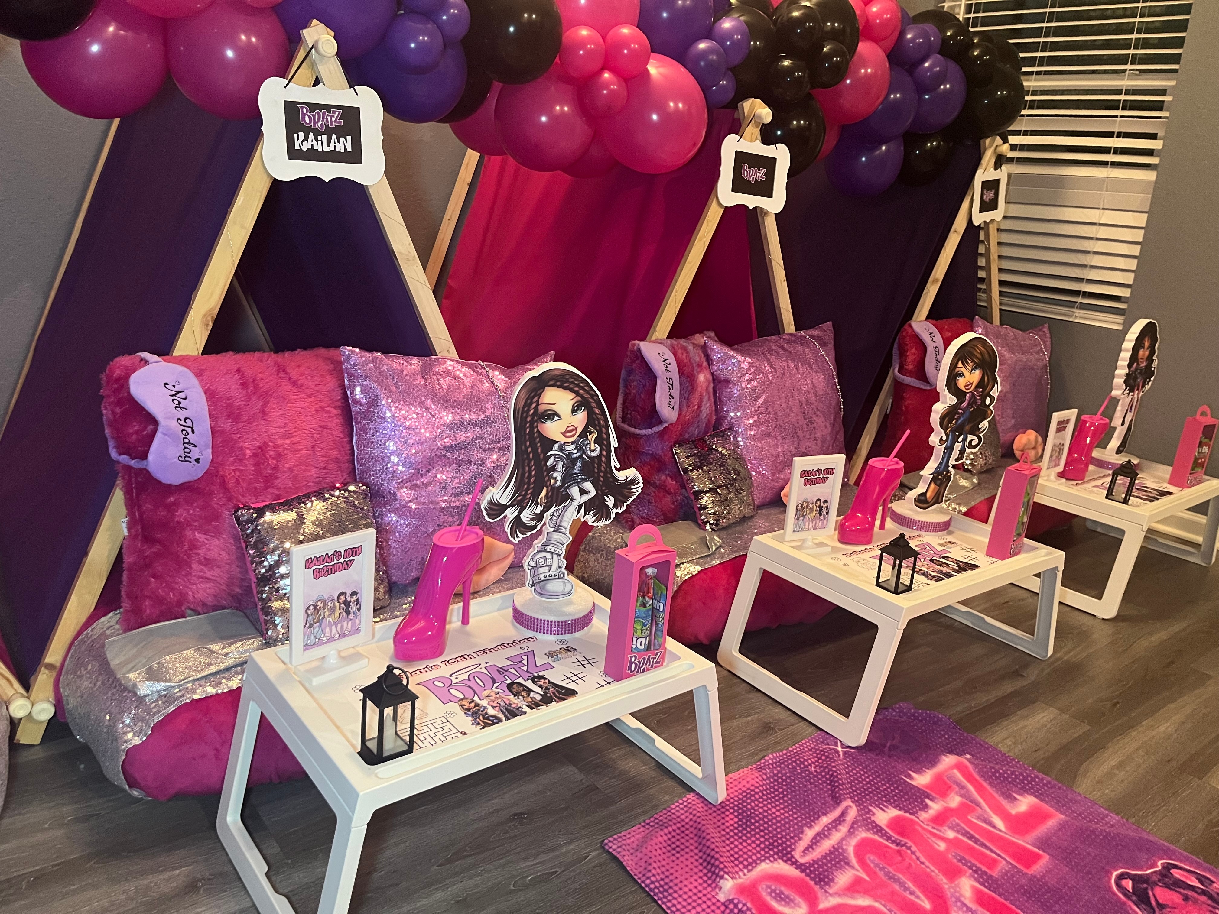 Party setup with pink, purple, and black balloons, decorated tables, and plush pillows with cartoon characters, pink bottles, and small tables.