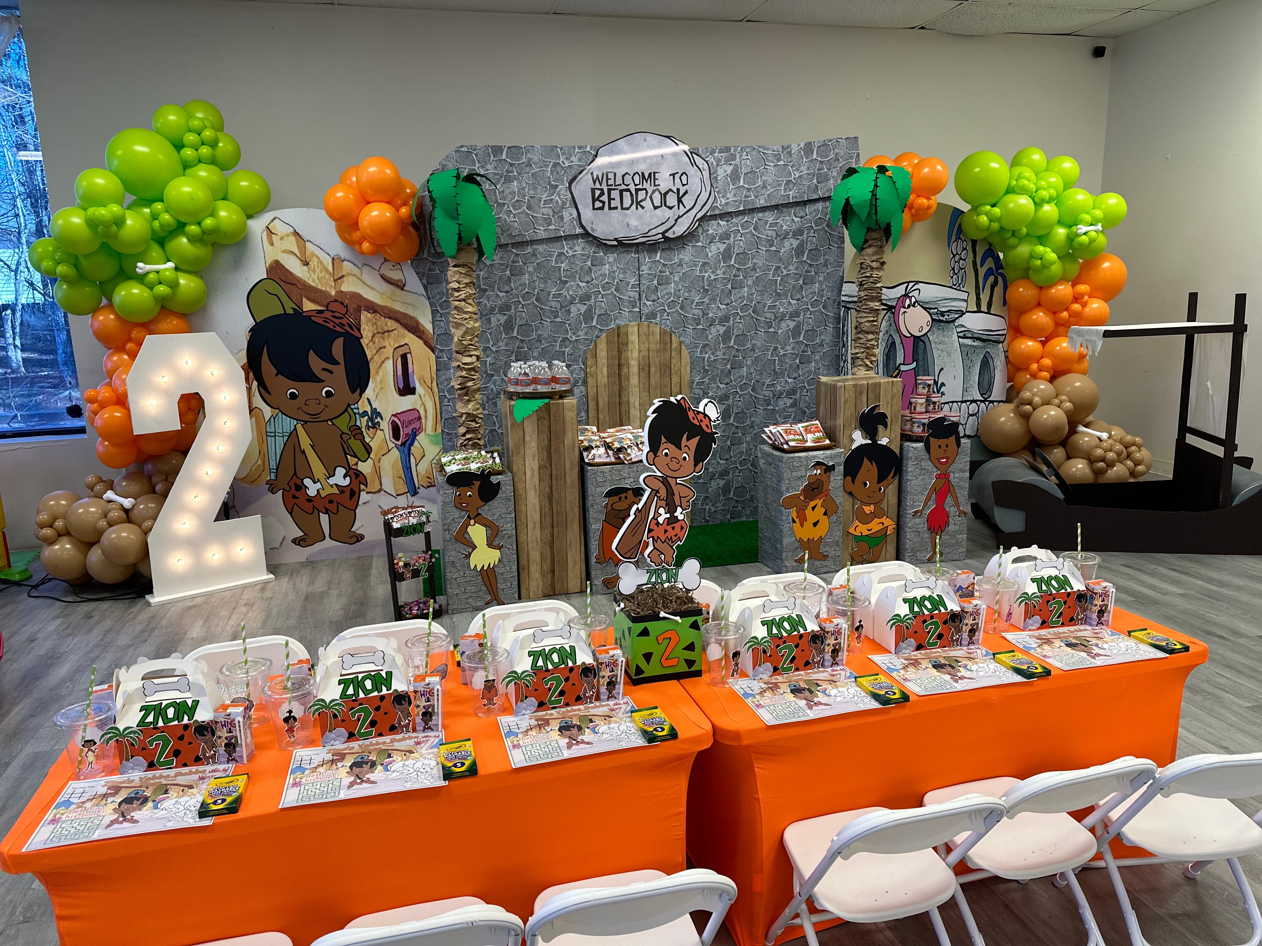 Decorative jungle-themed backdrop with children, balloons, and a table with party favors for a birthday celebration, with chairs in front.
