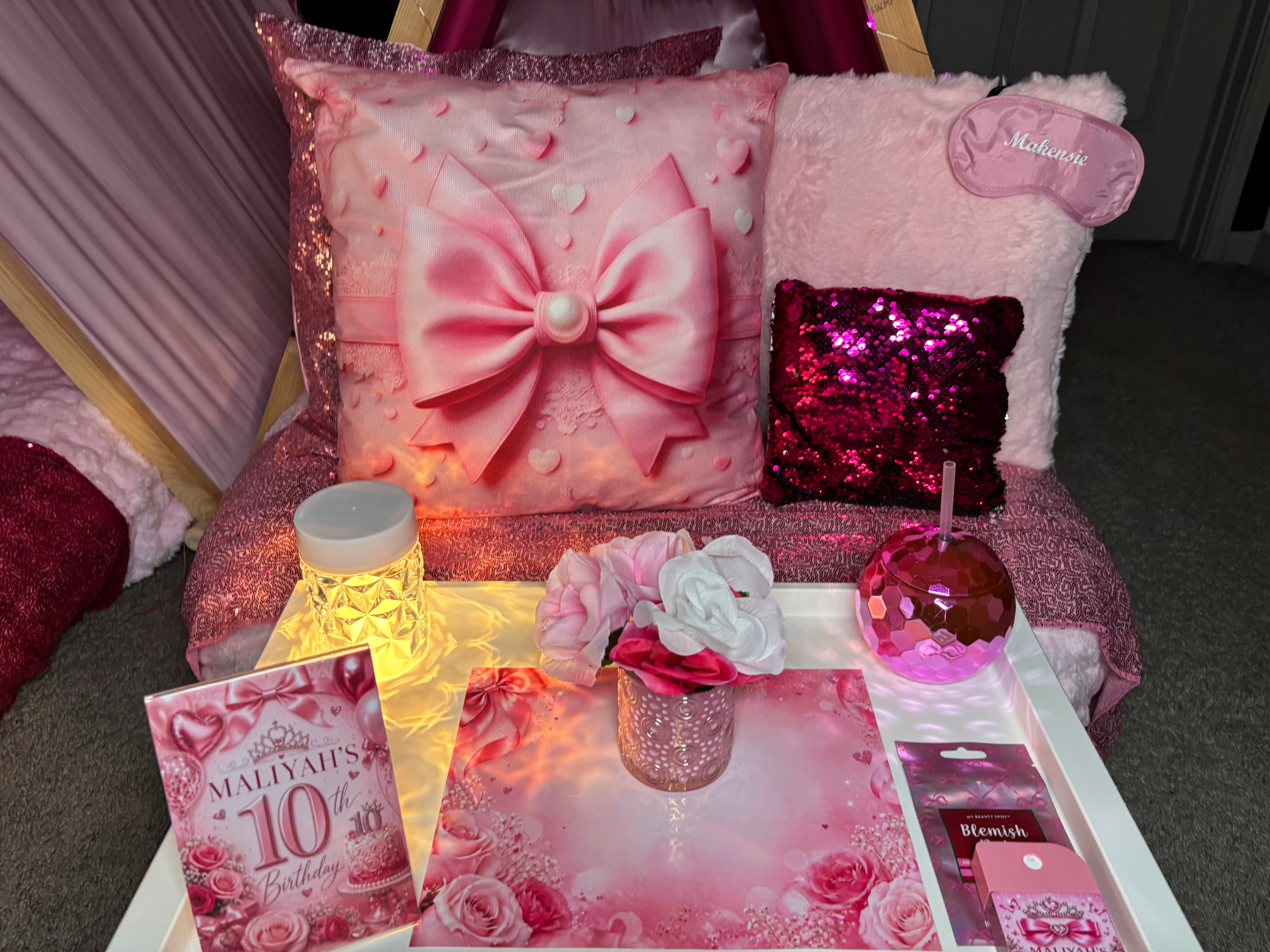 Decorative pillows on a pink surface, including a large pink pillow with a bow, a sequin pillow, and a plush pillow with a tag.