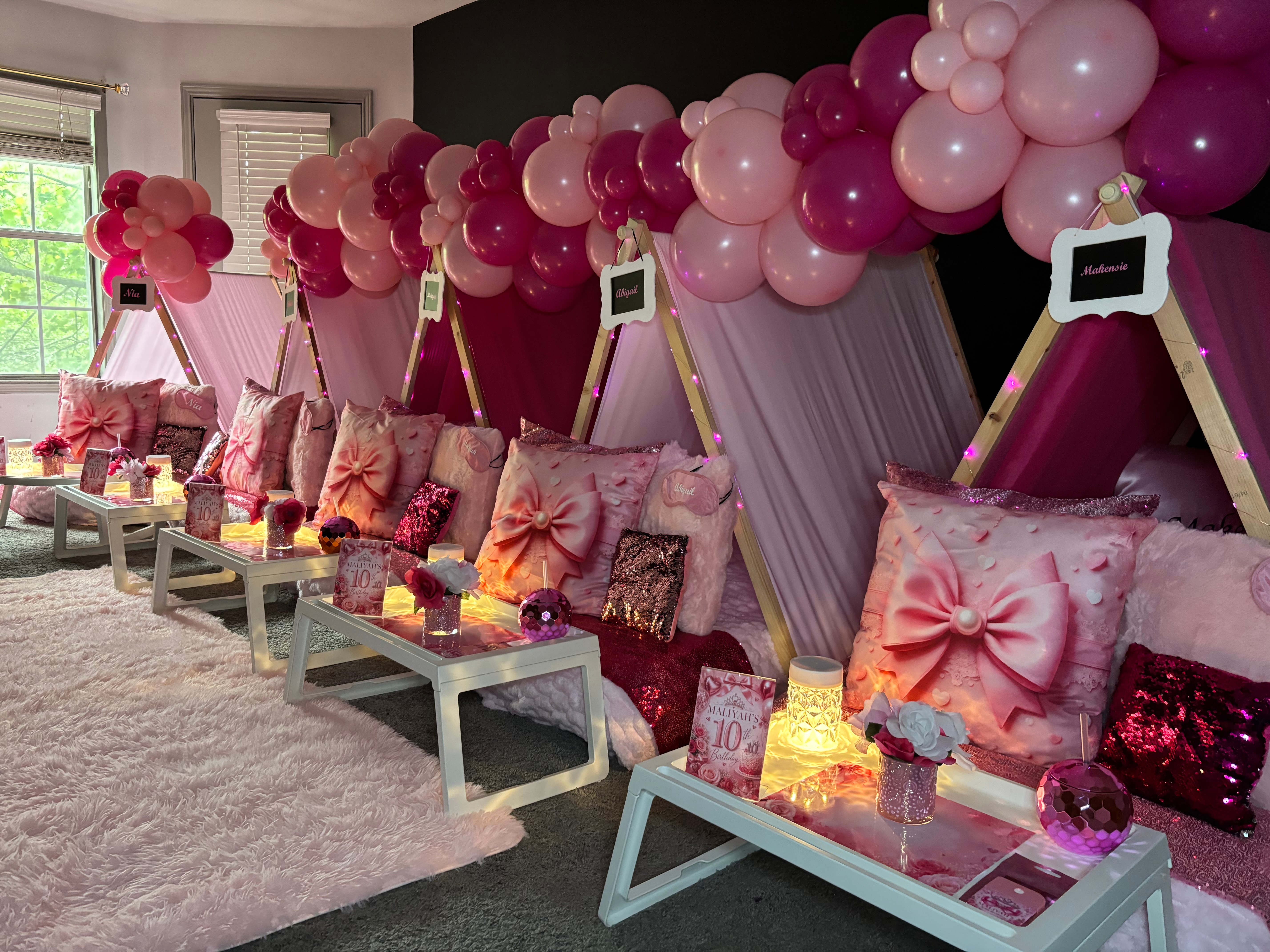 Decorated seating area with pink and purple balloons, pillows, and small tables, set against a pink backdrop with windows nearby.