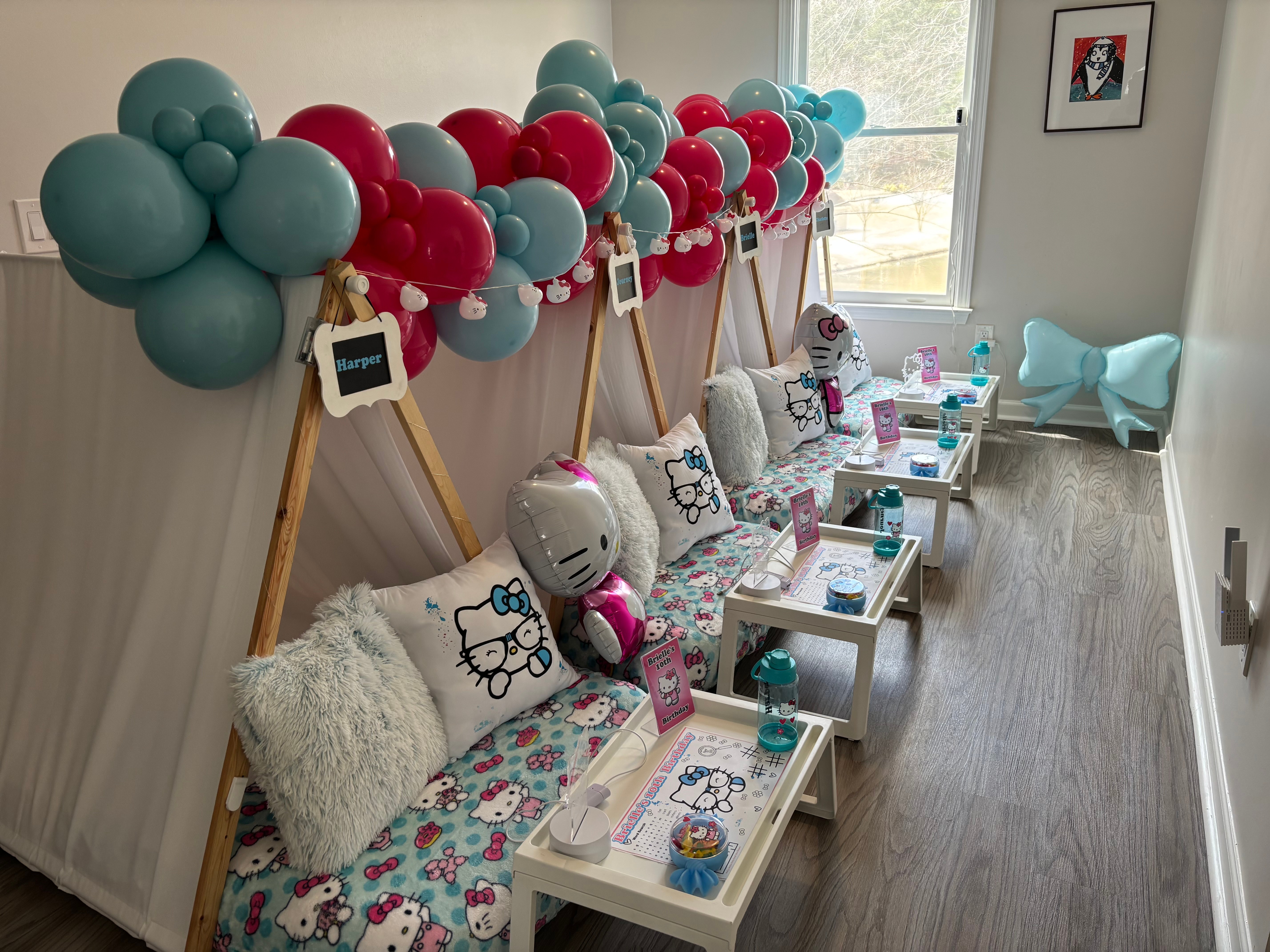Party setup with balloon decorations, small tables, and chairs along a wall near a window, with plush toys and party favors.