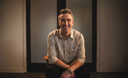 Smiling man with short hair and patterned shirt sitting in front of a dark door, arms crossed, in a warmly lit room.