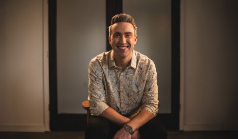 Smiling man with short hair and patterned shirt sitting in front of a dark door, arms crossed, in a warmly lit room.